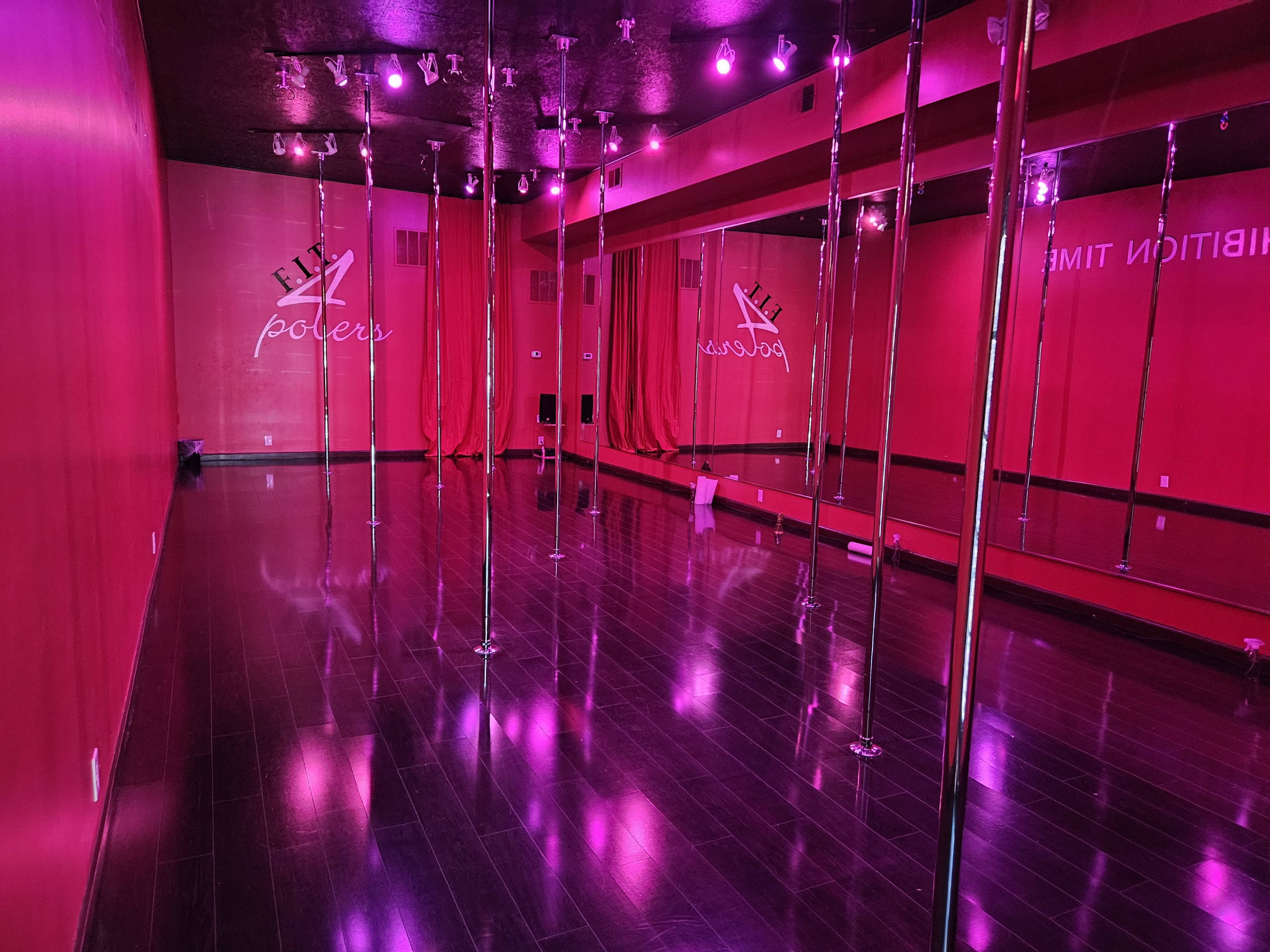 A dance studio with multiple pole dancing poles, mirrored walls, and pink lighting.
