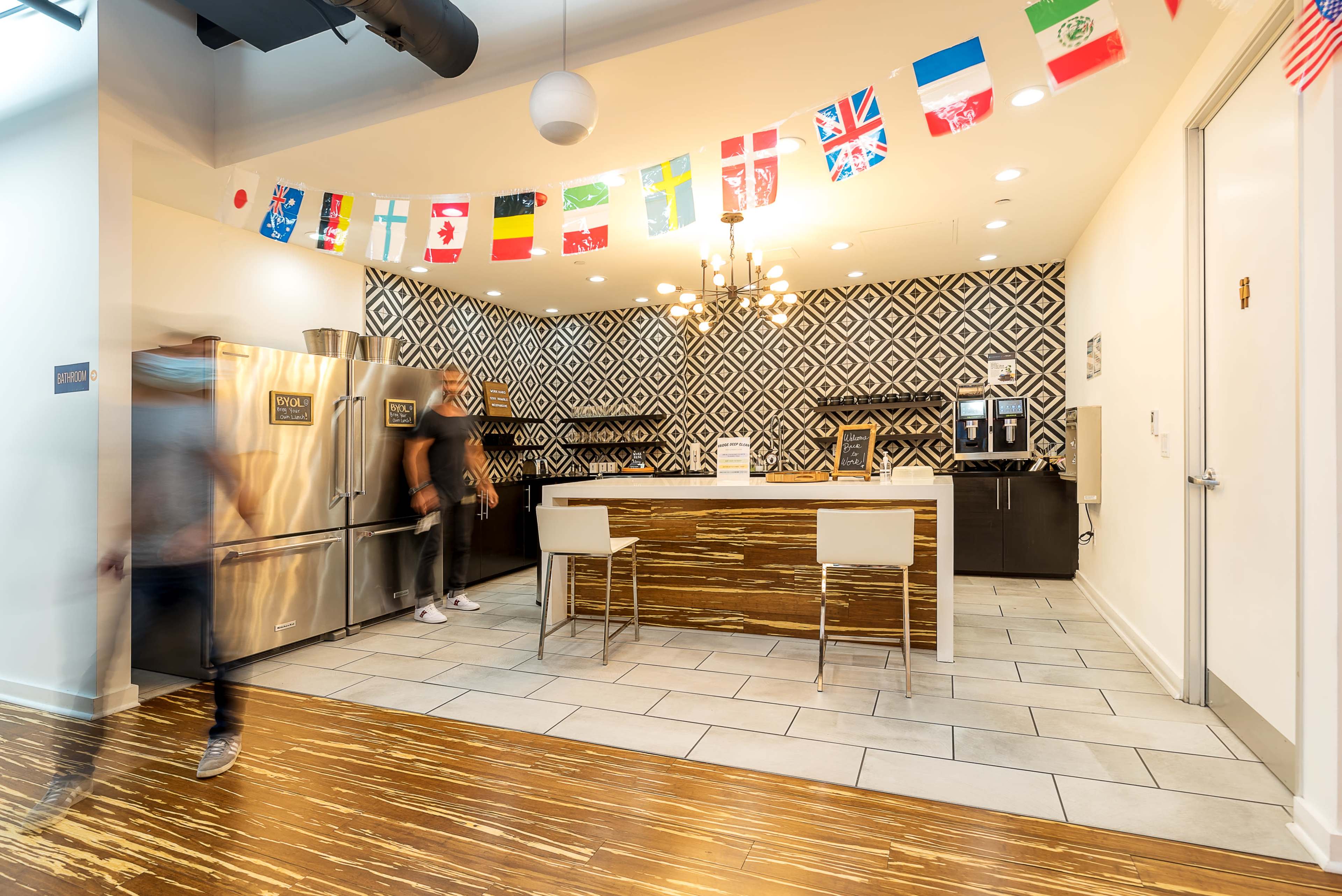 A modern kitchen area features a decorative wall with geometric patterns, bright flags hanging from the ceiling, and stainless steel appliances.
