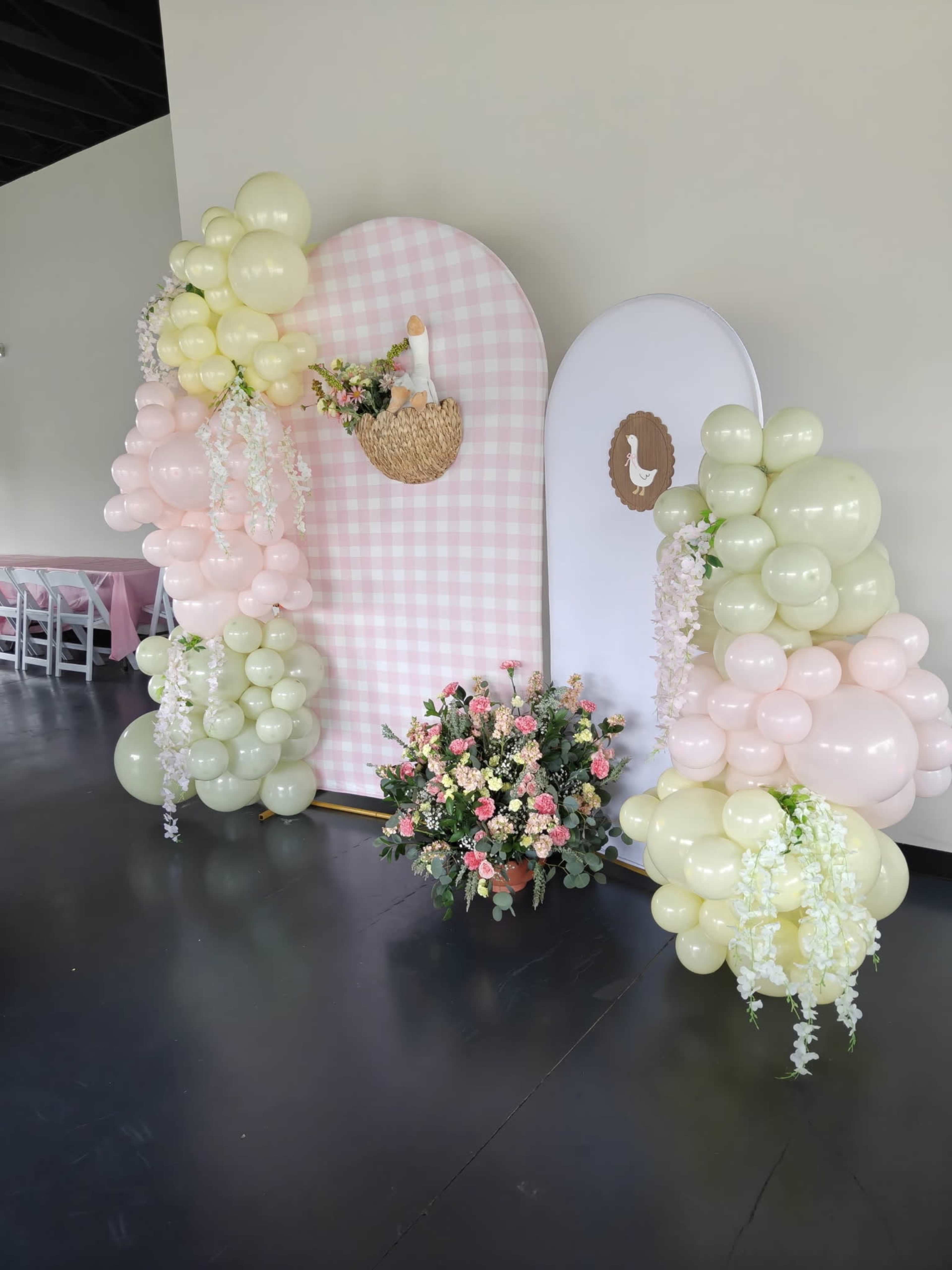The image shows a decorative setup featuring two large, rounded backdrops adorned with pastel-colored balloons and a floral arrangement at the base.