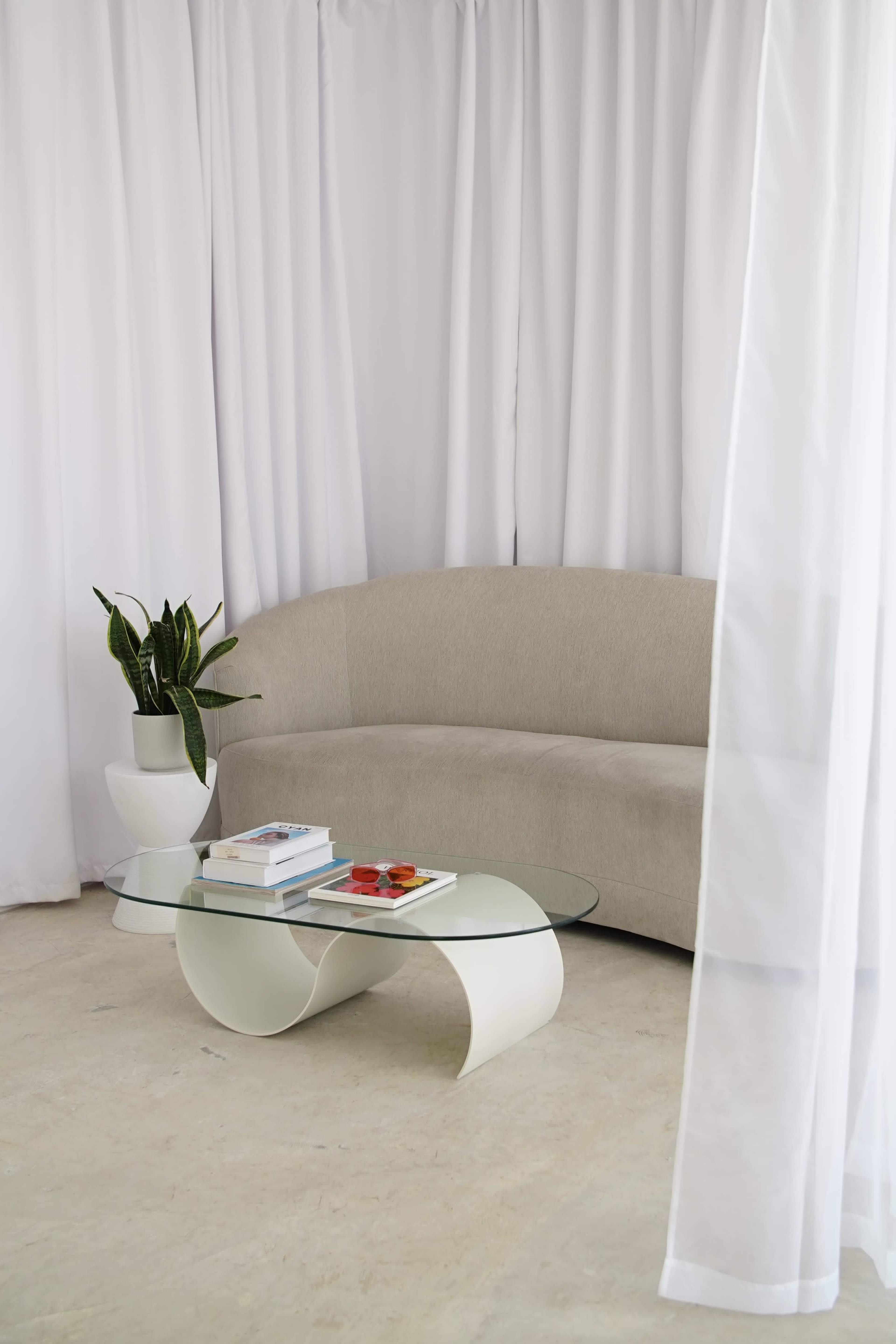 A curved beige sofa and a modern glass coffee table are positioned in front of sheer white curtains and a potted plant.