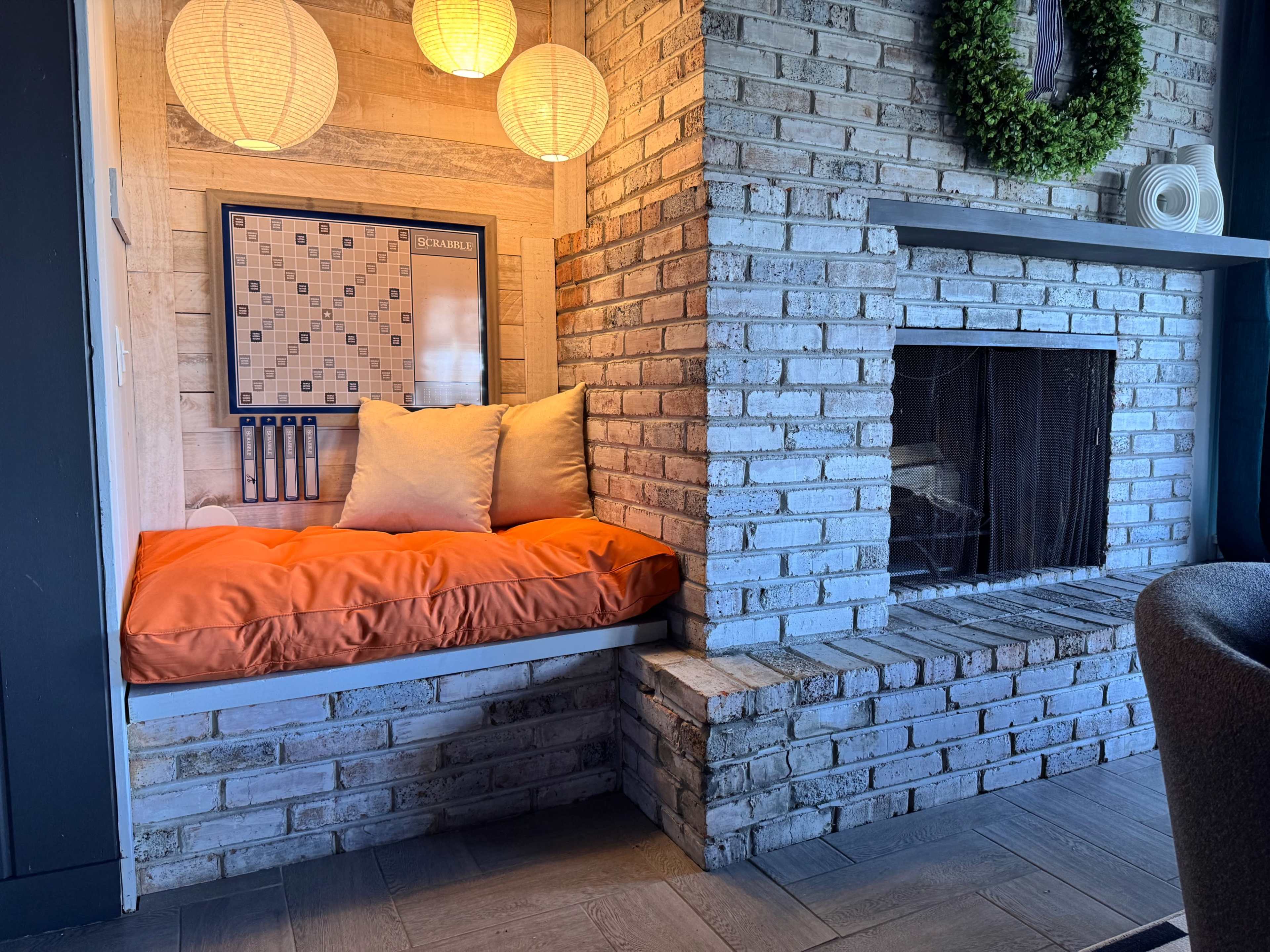 The image shows a cozy nook with an orange cushion built into a brick wall next to a fireplace, illuminated by two hanging lanterns.