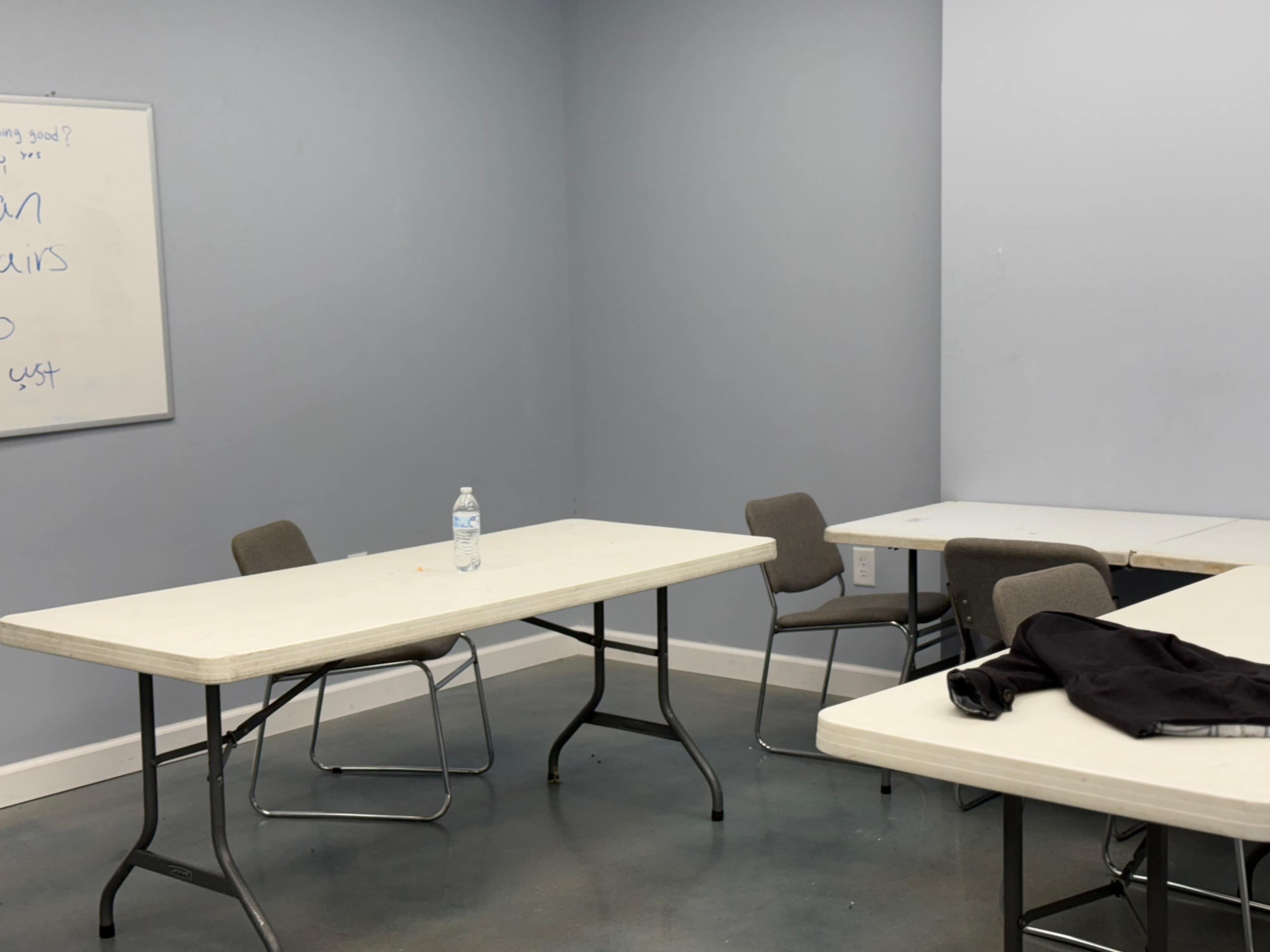 The image shows a sparse room with two empty tables and several chairs, along with a water bottle on one of the tables and a black garment draped over another.