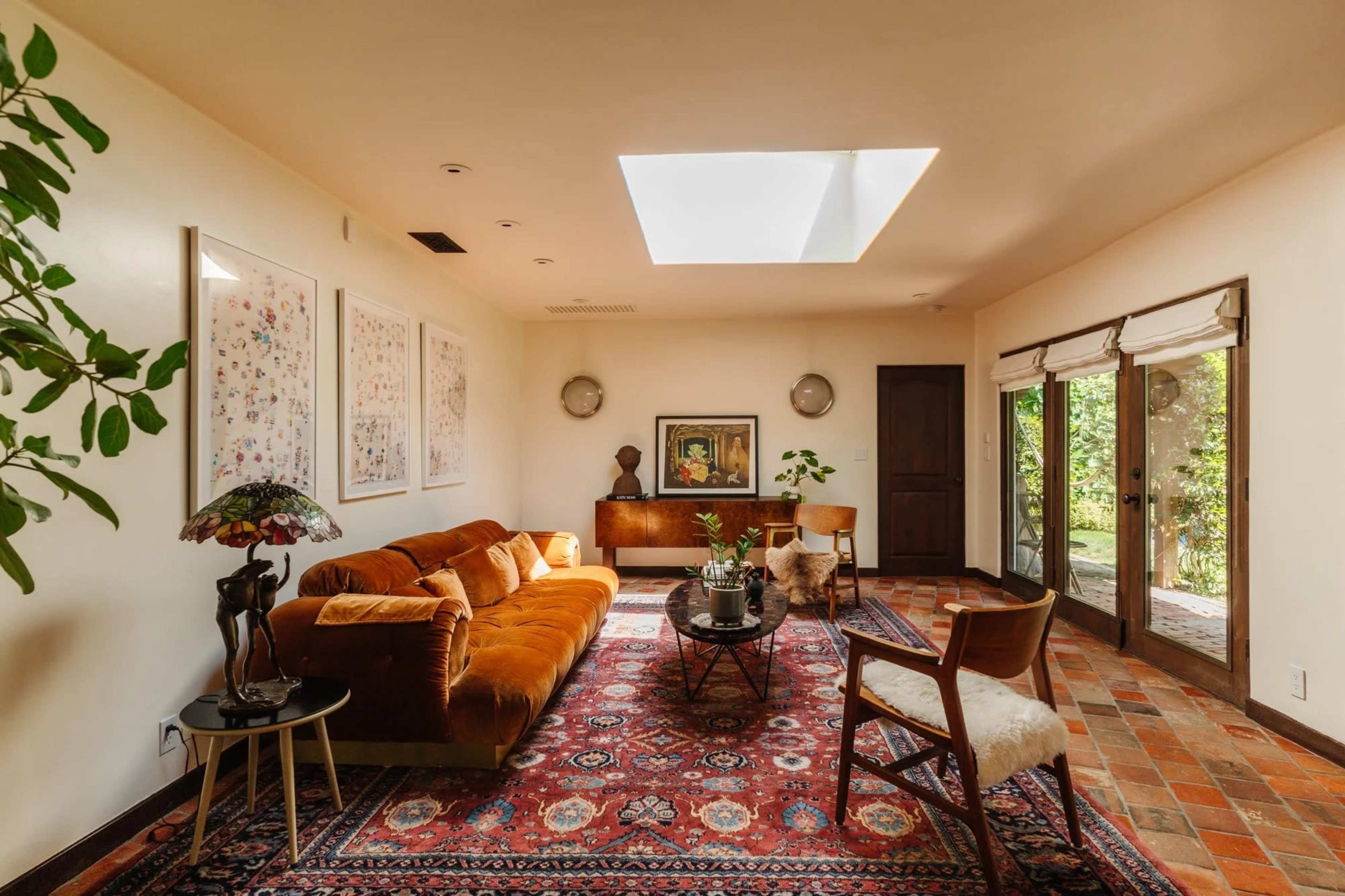 The room features a brown sofa, a coffee table, and large glass doors that open to a garden, illuminated by a skylight above.