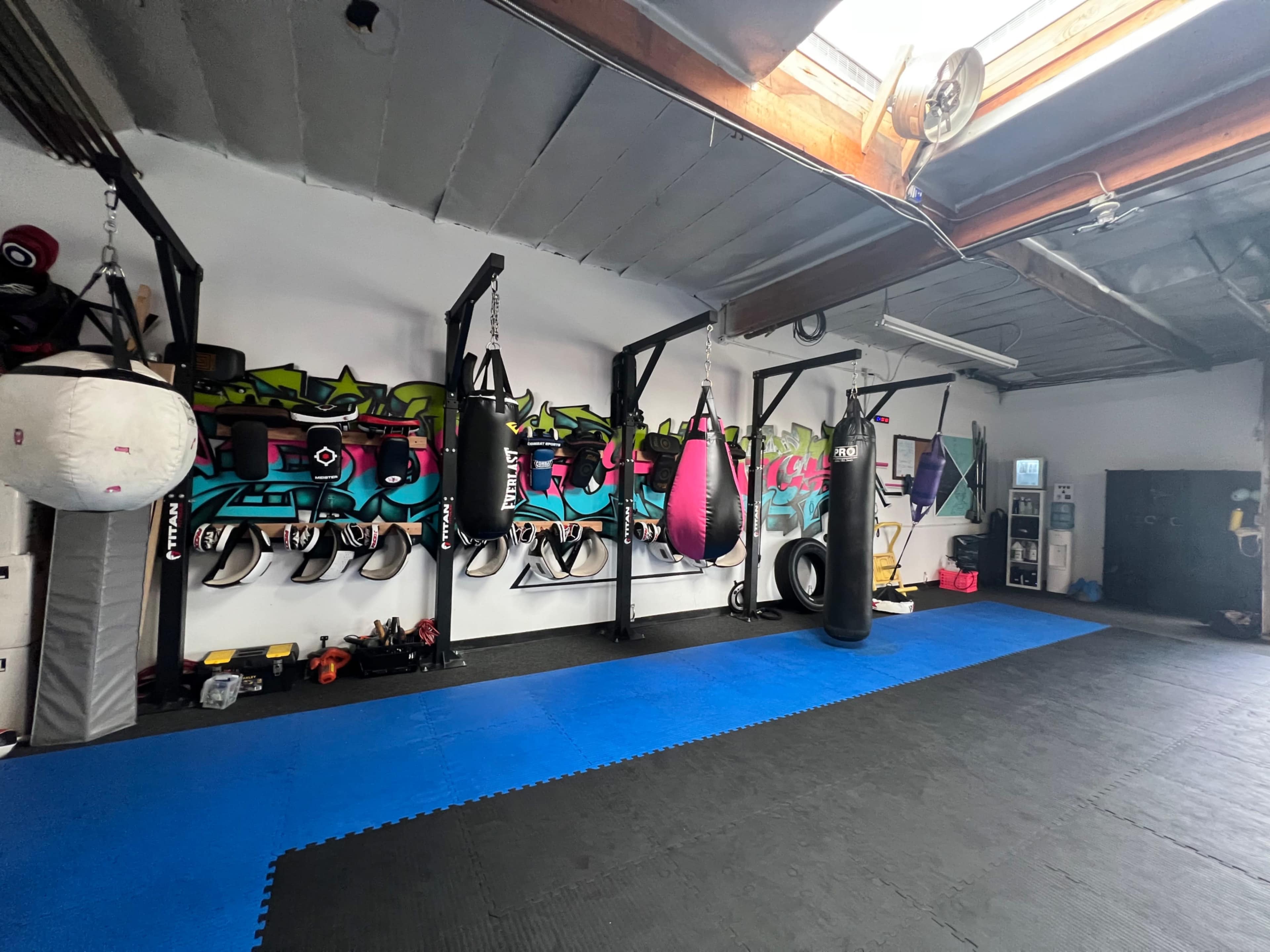 A boxing gym with several hanging punching bags, a padded blue floor, and a backdrop of colorful graffiti on the wall.