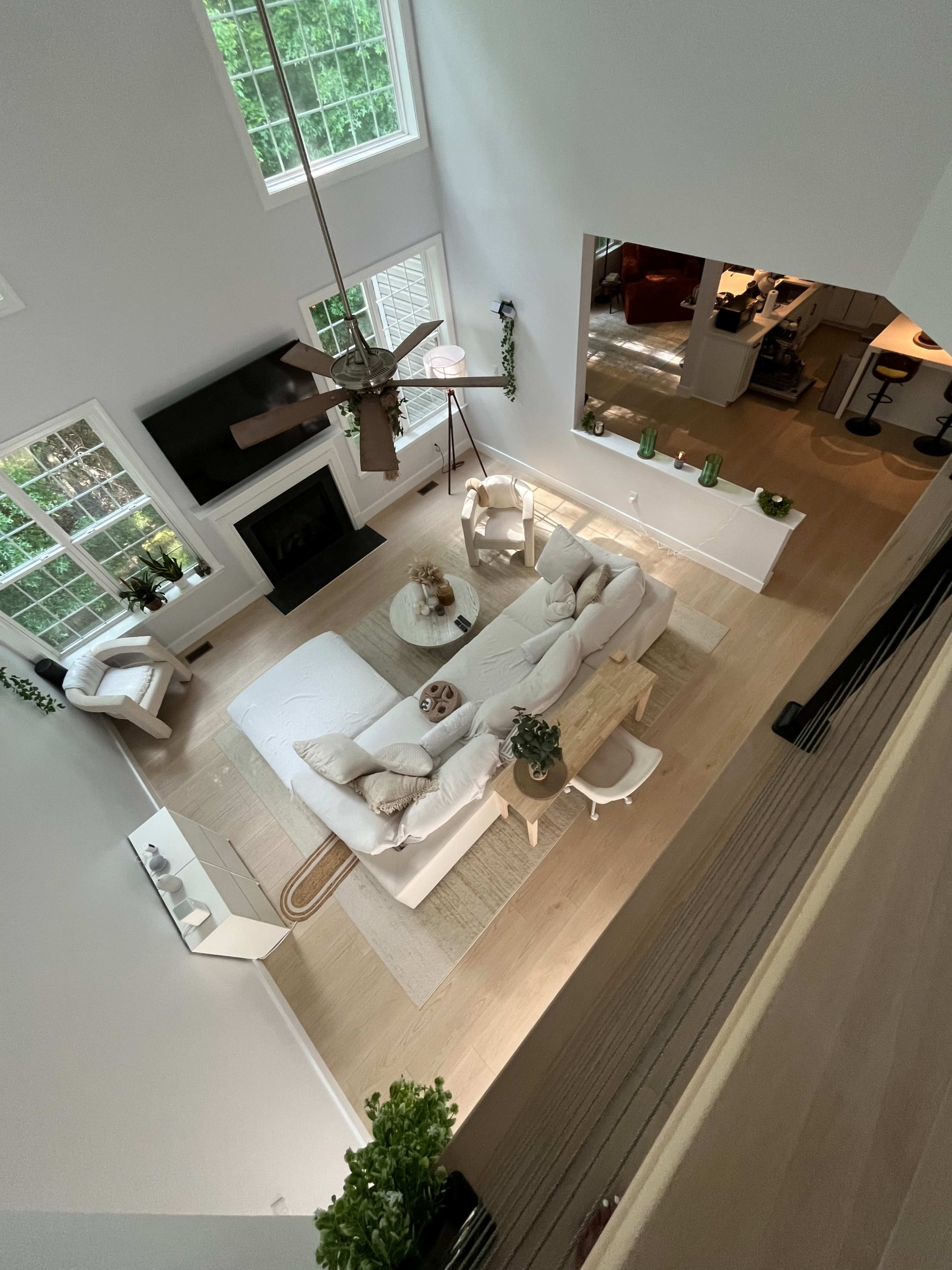 The image shows a spacious, well-lit living room with a white sofa, a coffee table, and a fireplace, surrounded by large windows and minimalistic decor.