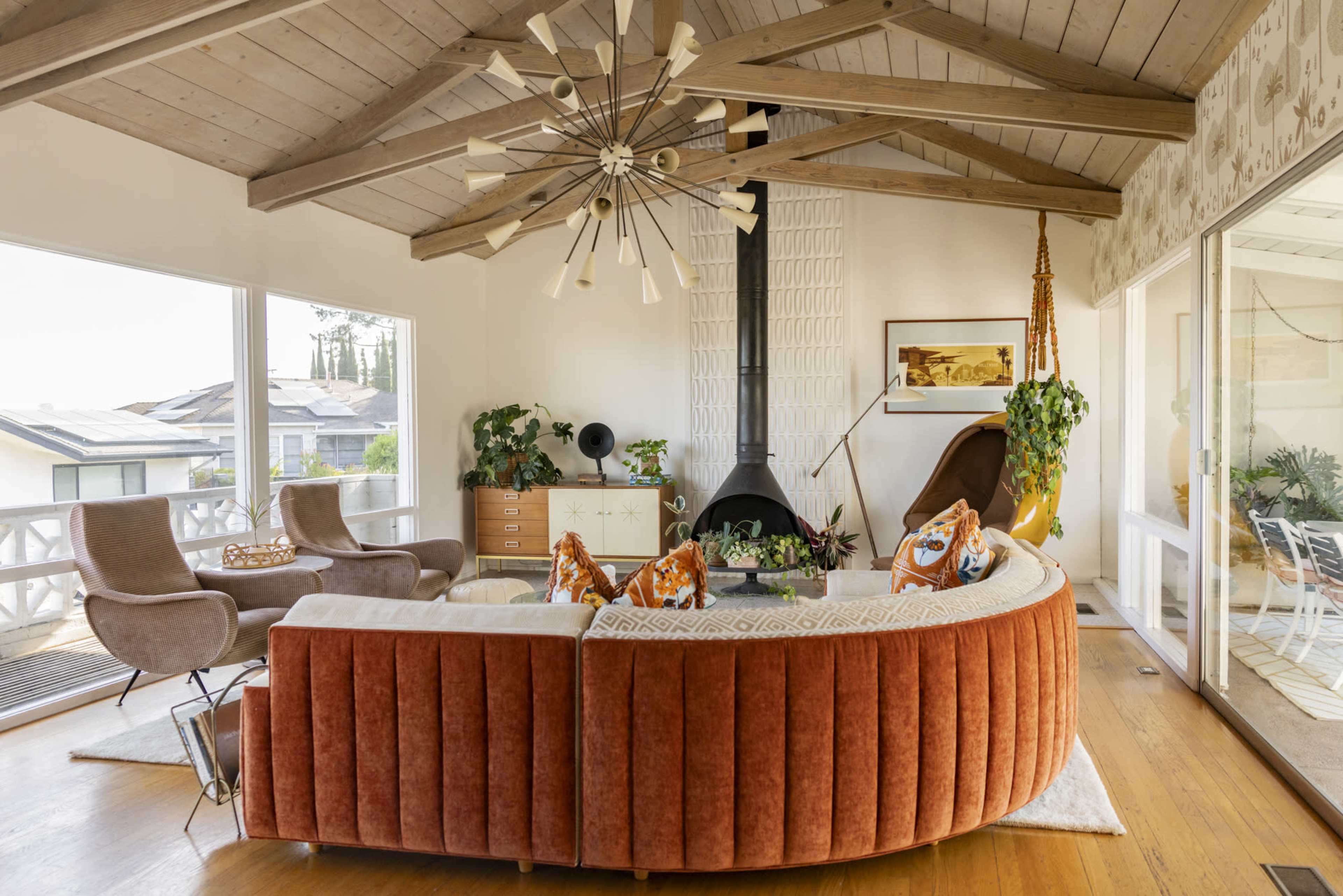 The image shows a modern living room with a curved orange sofa, two brown armchairs, a large window, and a geometric ceiling light fixture.