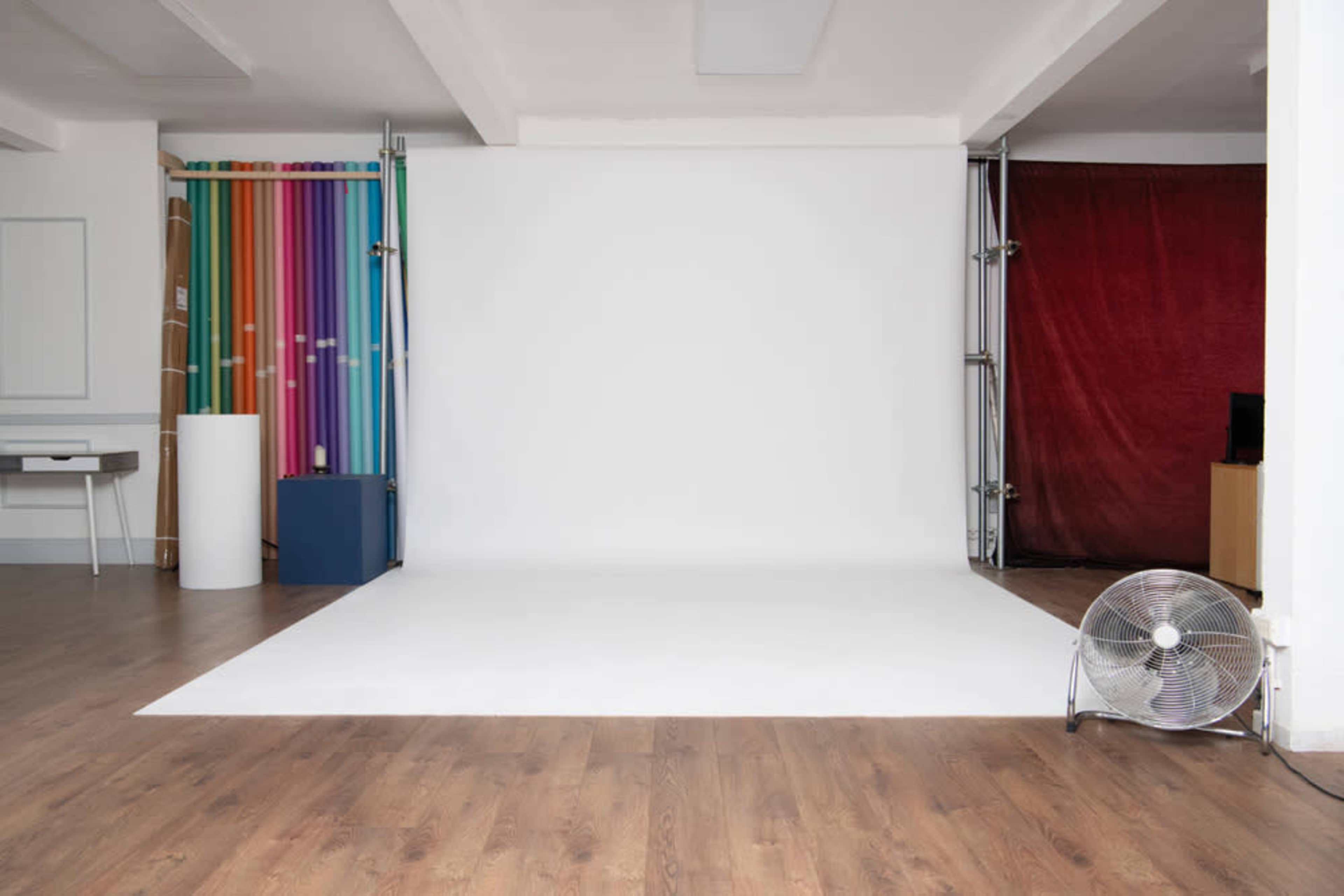 The image shows a photography studio with a blank white backdrop and colorful striped rolls on one side, along with a fan and wooden flooring.