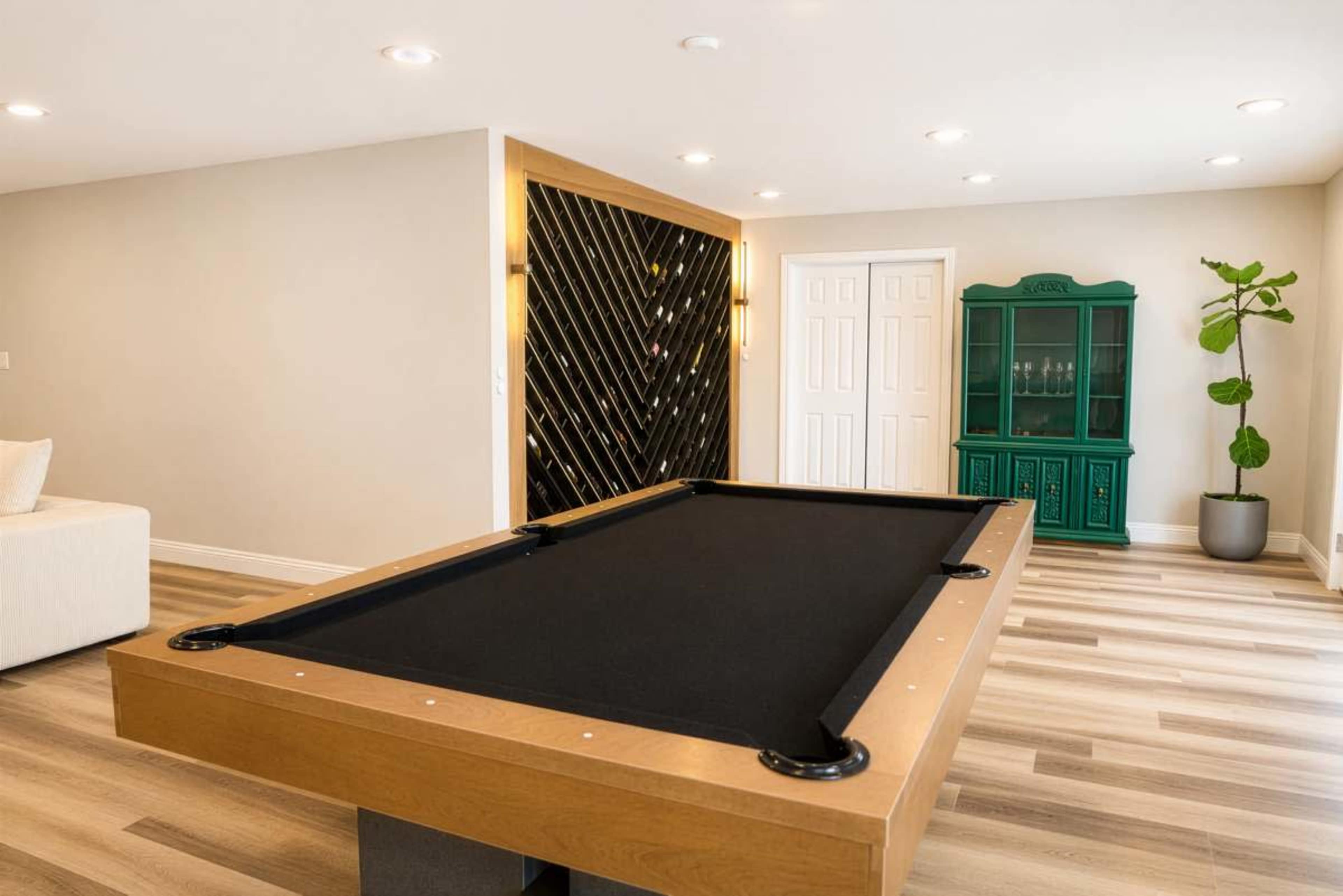 The image shows a modern game room featuring a sleek pool table with a black felt surface, wooden edges, and a green cabinet against the wall.