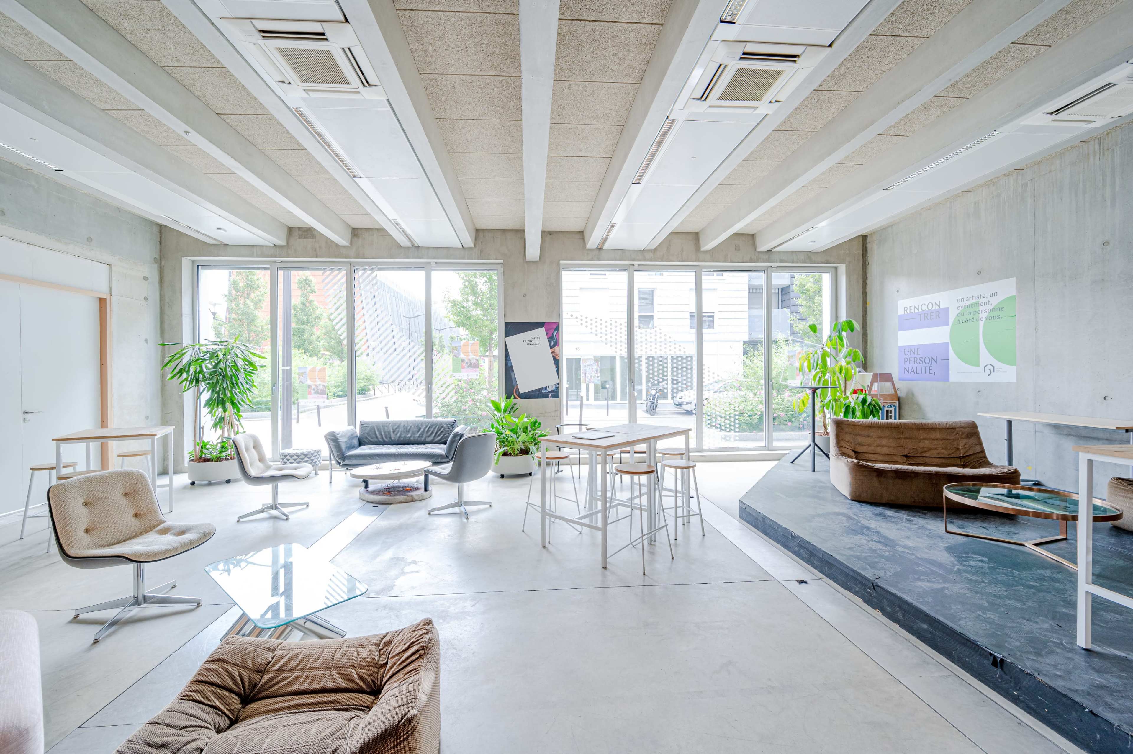 A modern workspace with concrete walls, large windows letting in natural light, and a mix of seating arrangements including chairs, tables, and plants.
