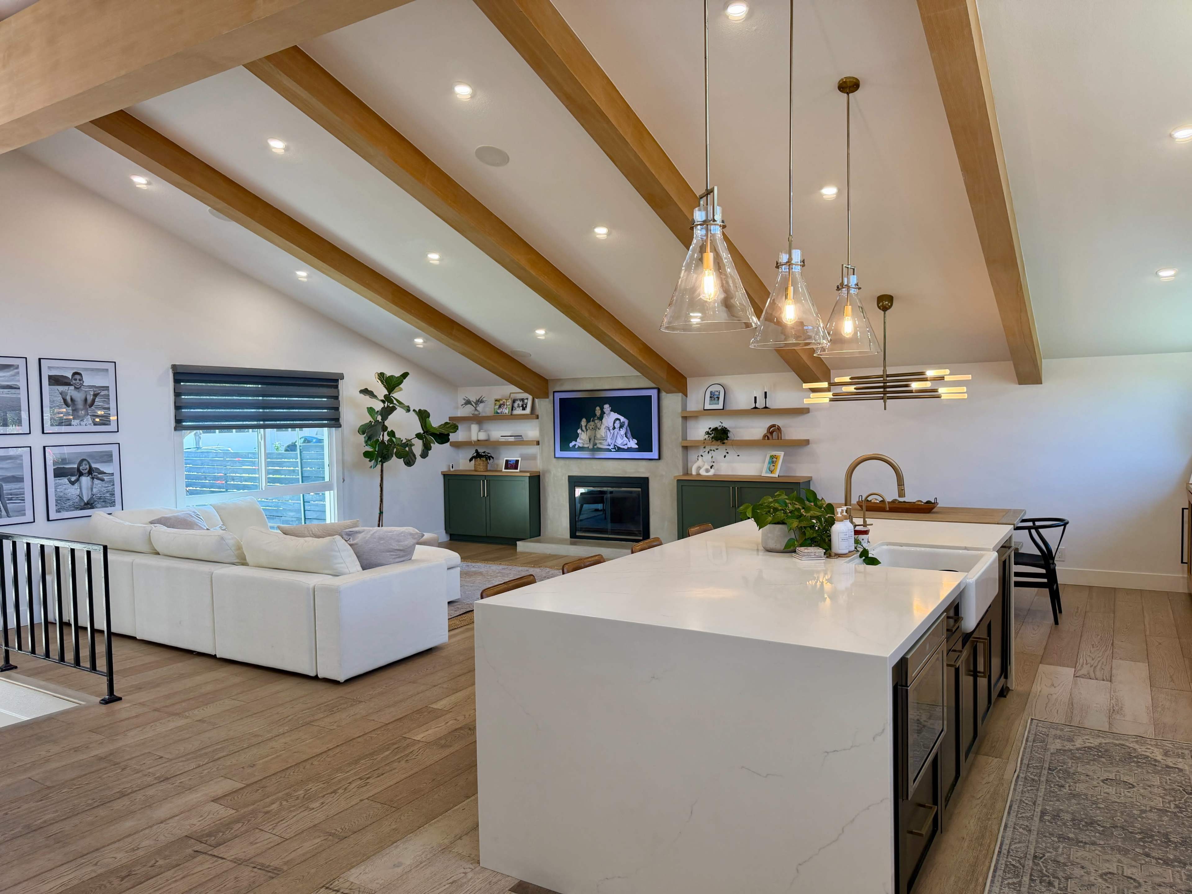 The image shows a modern living space with a high, beamed ceiling, featuring a white sectional sofa, a large television mounted on the wall, and a kitchen island with pendant lighting and a sink.