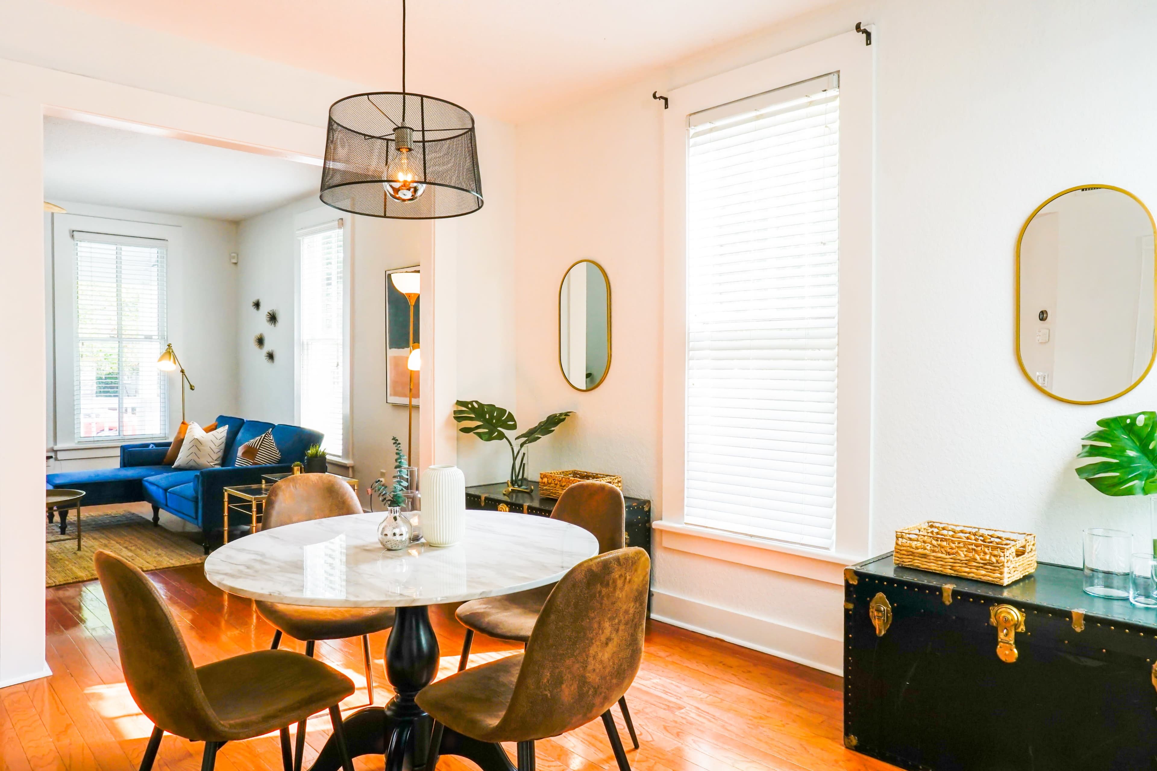 A bright and airy dining area featuring a round marble table surrounded by brown chairs, with a blue sofa and decorative plants visible in the background.