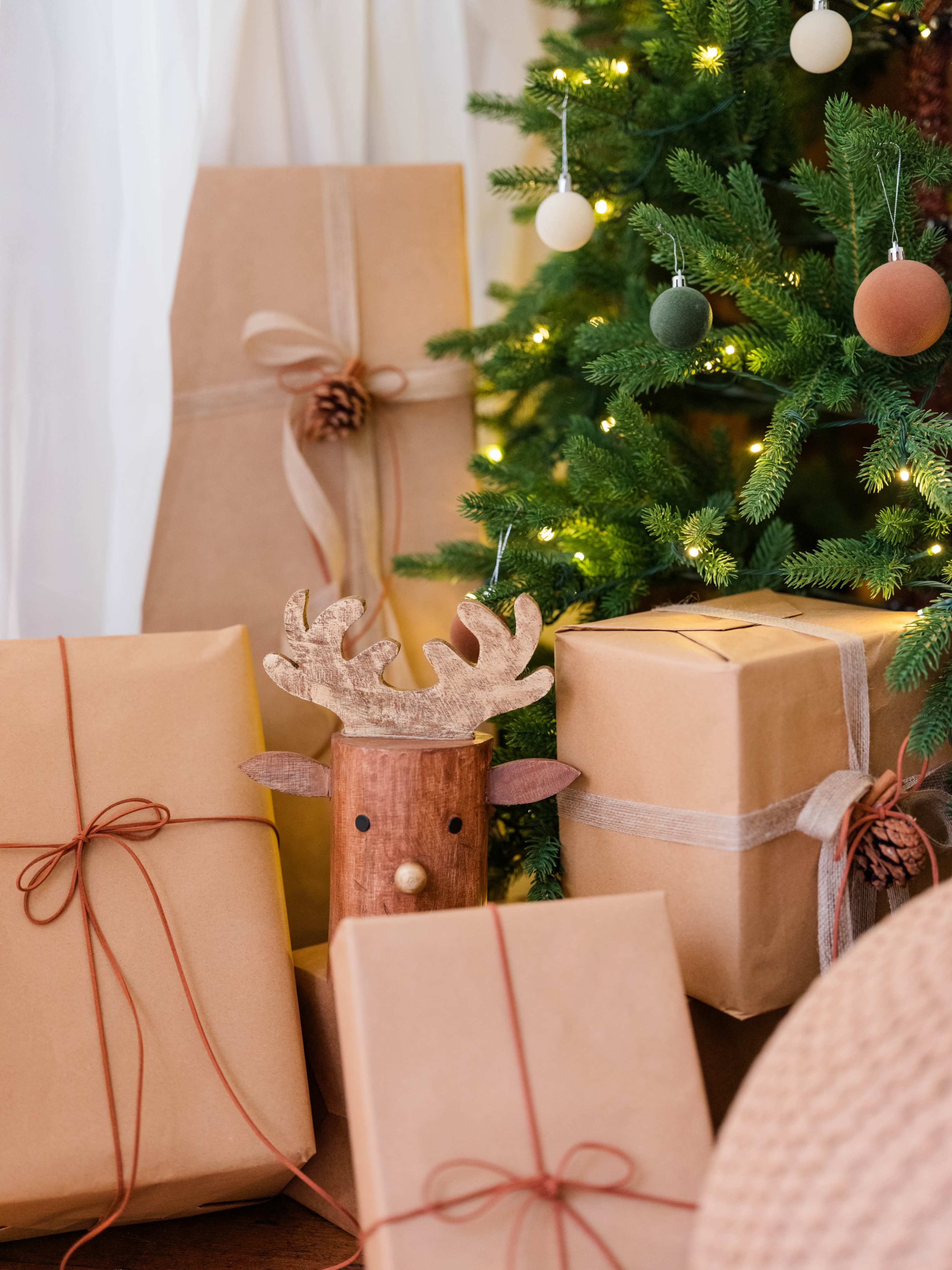 The image shows a collection of wrapped gifts in brown paper, with a wooden reindeer decoration, arranged near a decorated Christmas tree.