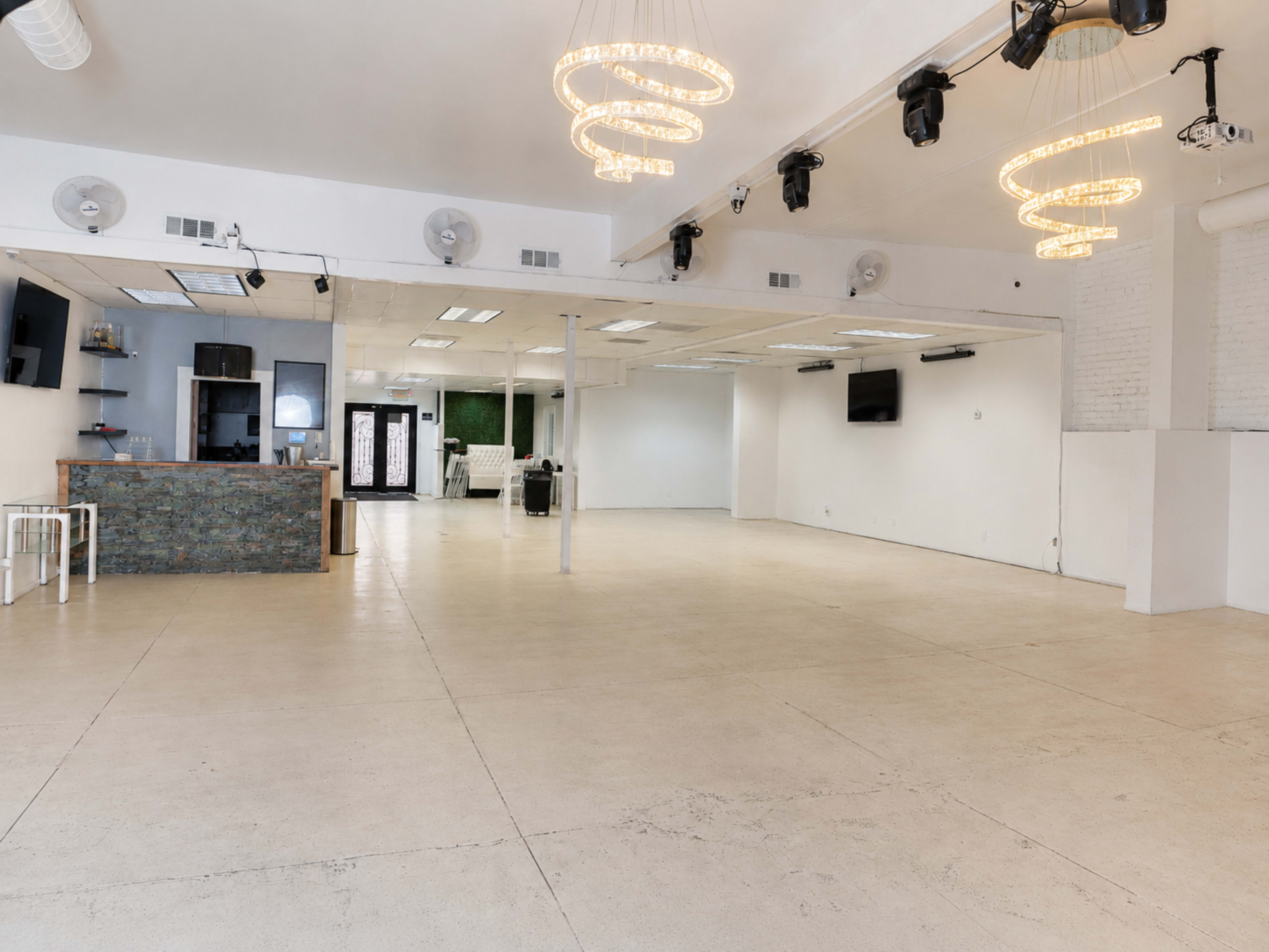 The image shows a spacious, empty indoor venue with white walls, concrete flooring, and decorative light fixtures hanging from the ceiling.