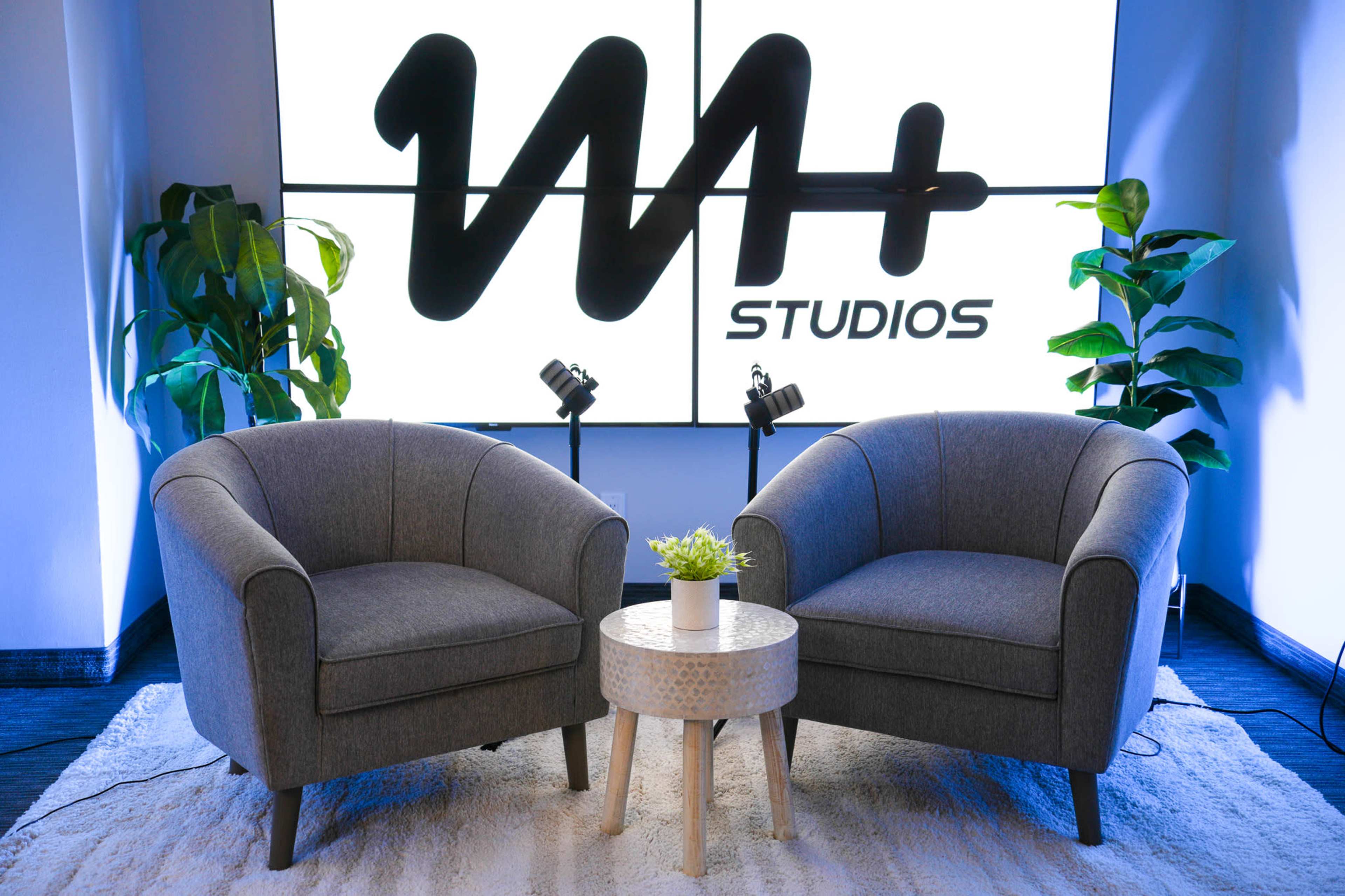 The image shows a pair of gray armchairs positioned around a small circular table with a potted plant, set against a large wall displaying a logo for "W+ STUDIOS."