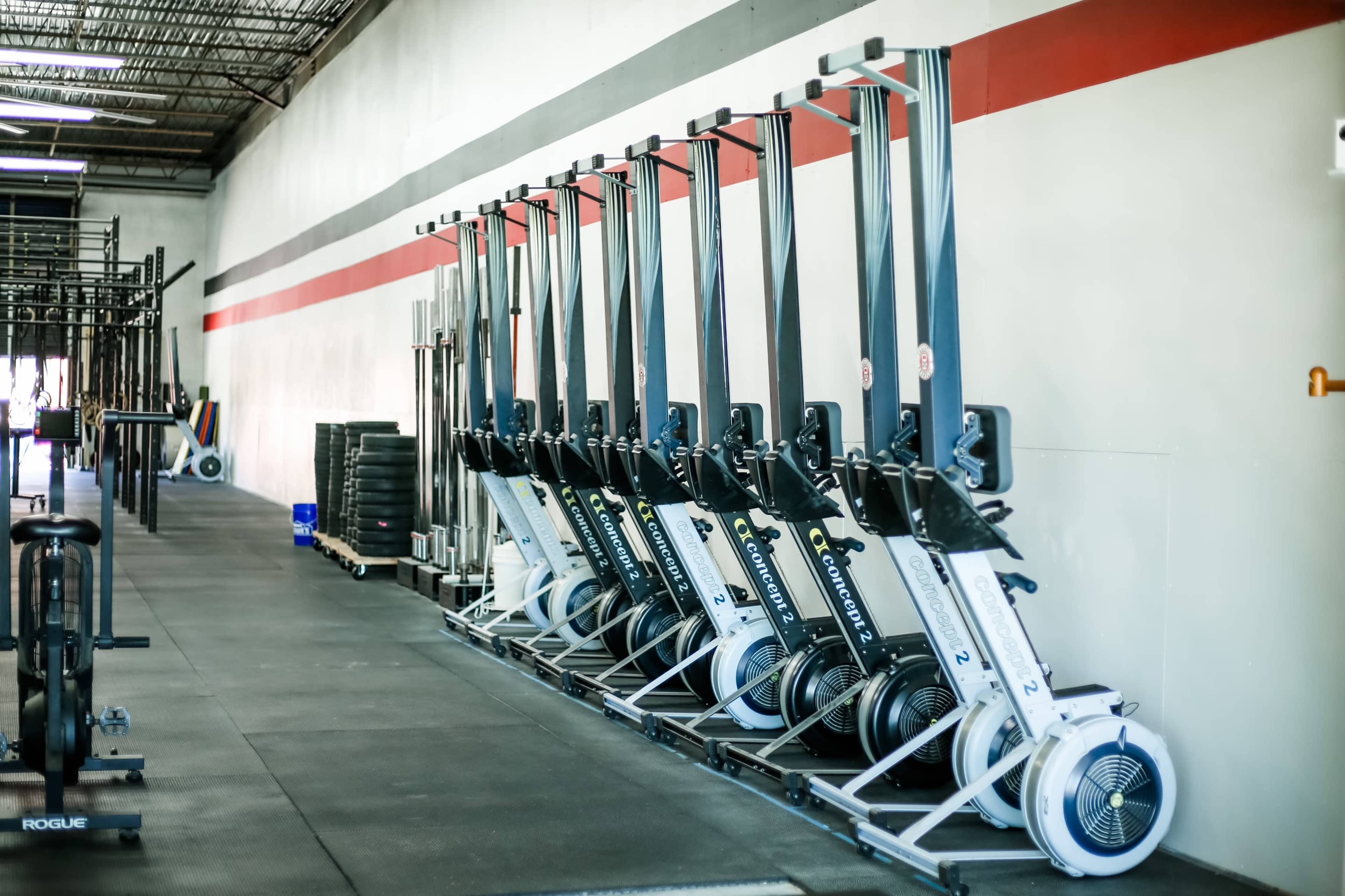 A row of rowing machines lined up against a warehouse-style gym wall with red and white paint.