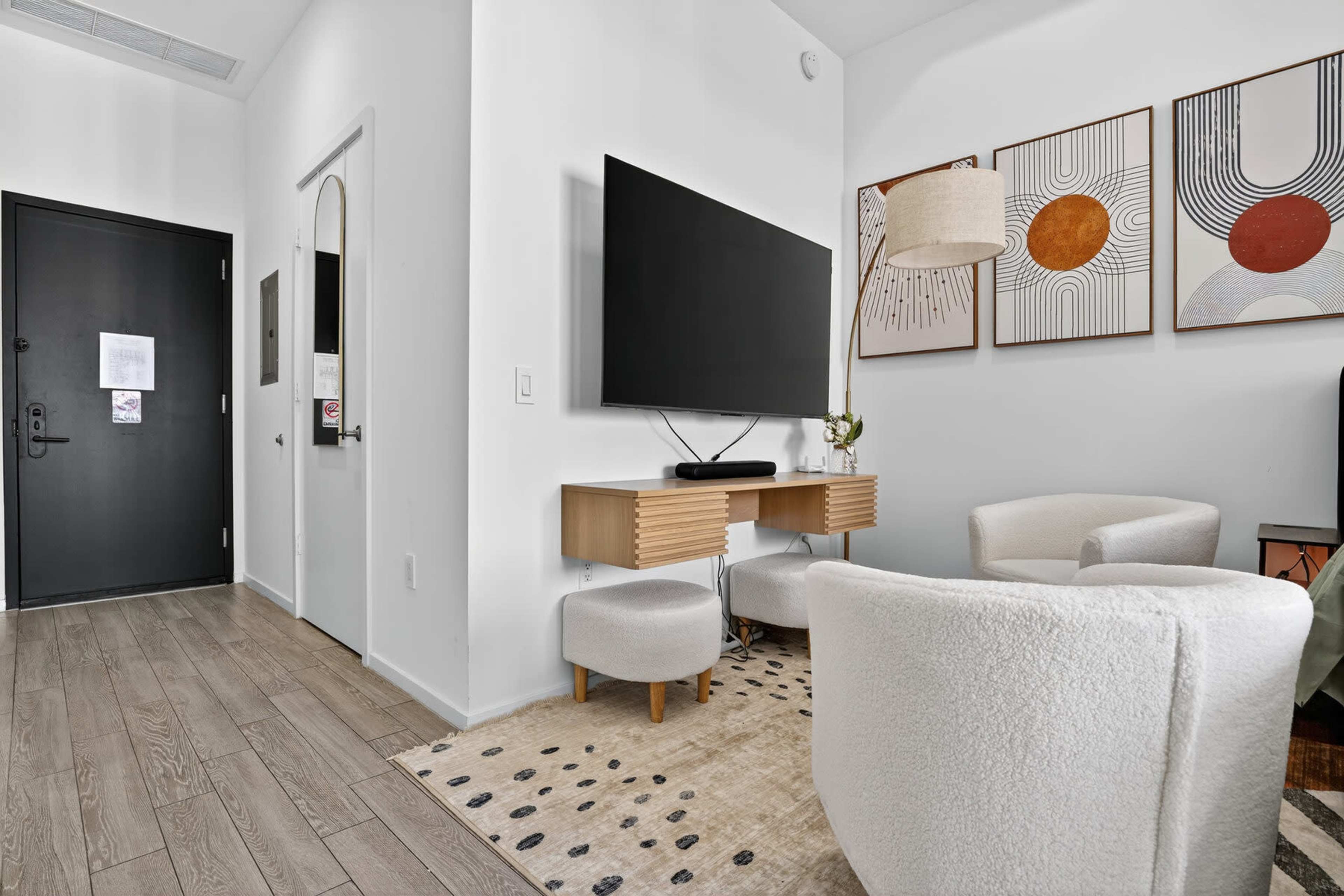 The image shows a modern living space featuring a small seating area with two light chairs, a wall-mounted TV, a wooden console, and abstract artwork on the walls.