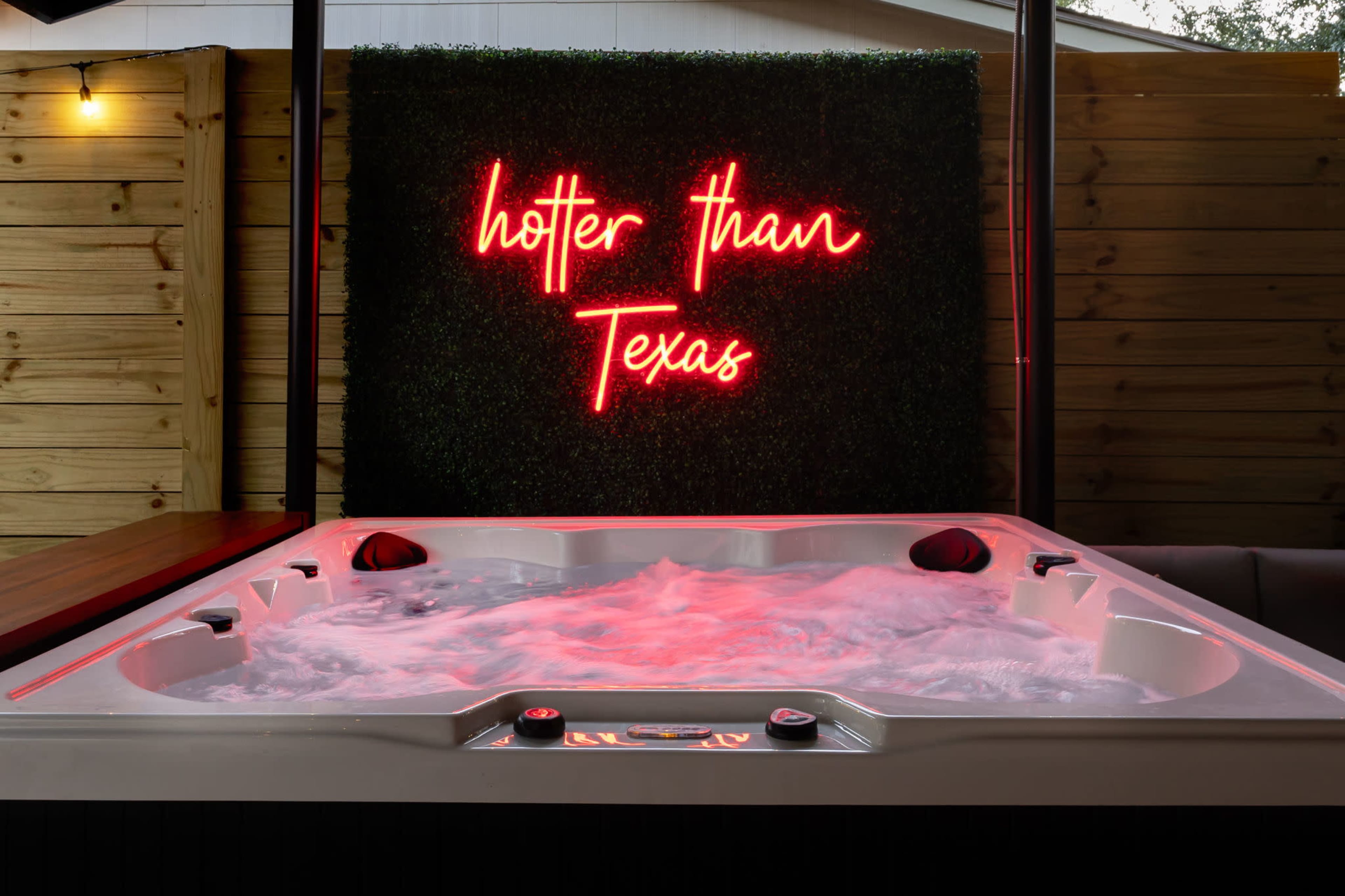 A hot tub with bubbling water is illuminated by a neon sign that reads "hotter than Texas" against a green wall.