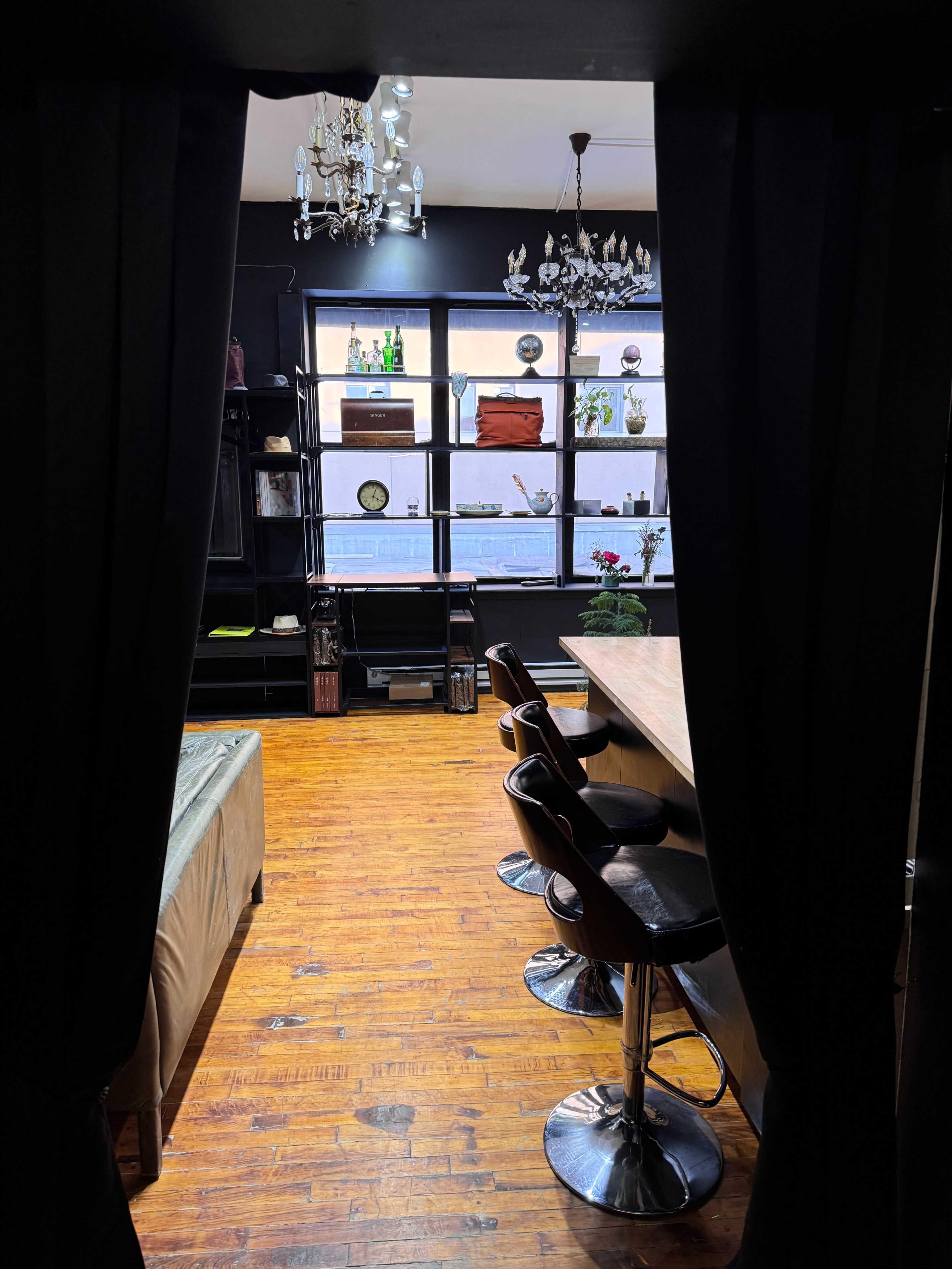 A view from behind a curtain into a room featuring a wooden floor, a long table with bar stools, and shelves displaying various items against a dark wall.