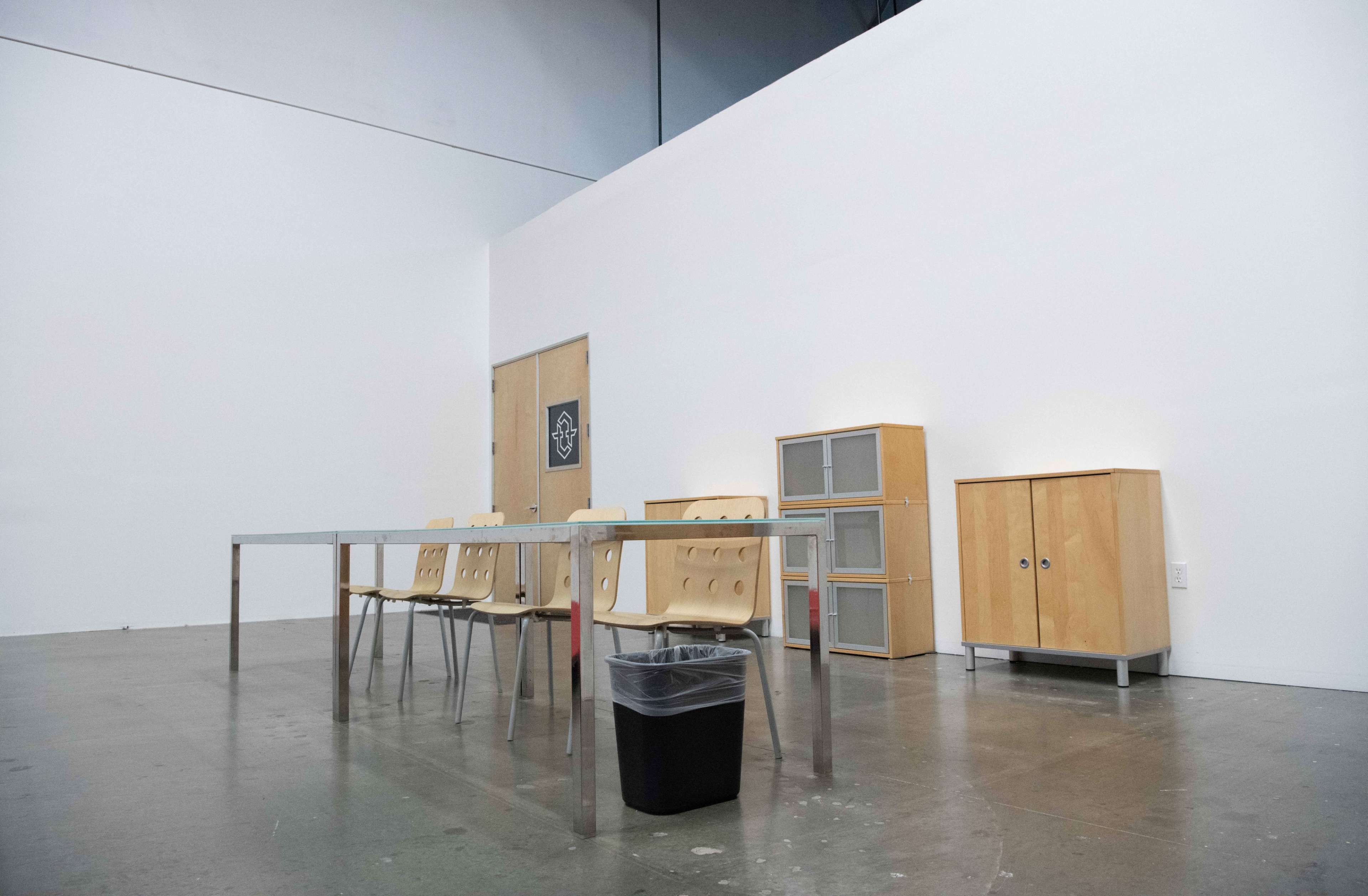 An empty room features a long table with four chairs, a trash bin underneath, and wooden cabinets against the wall.