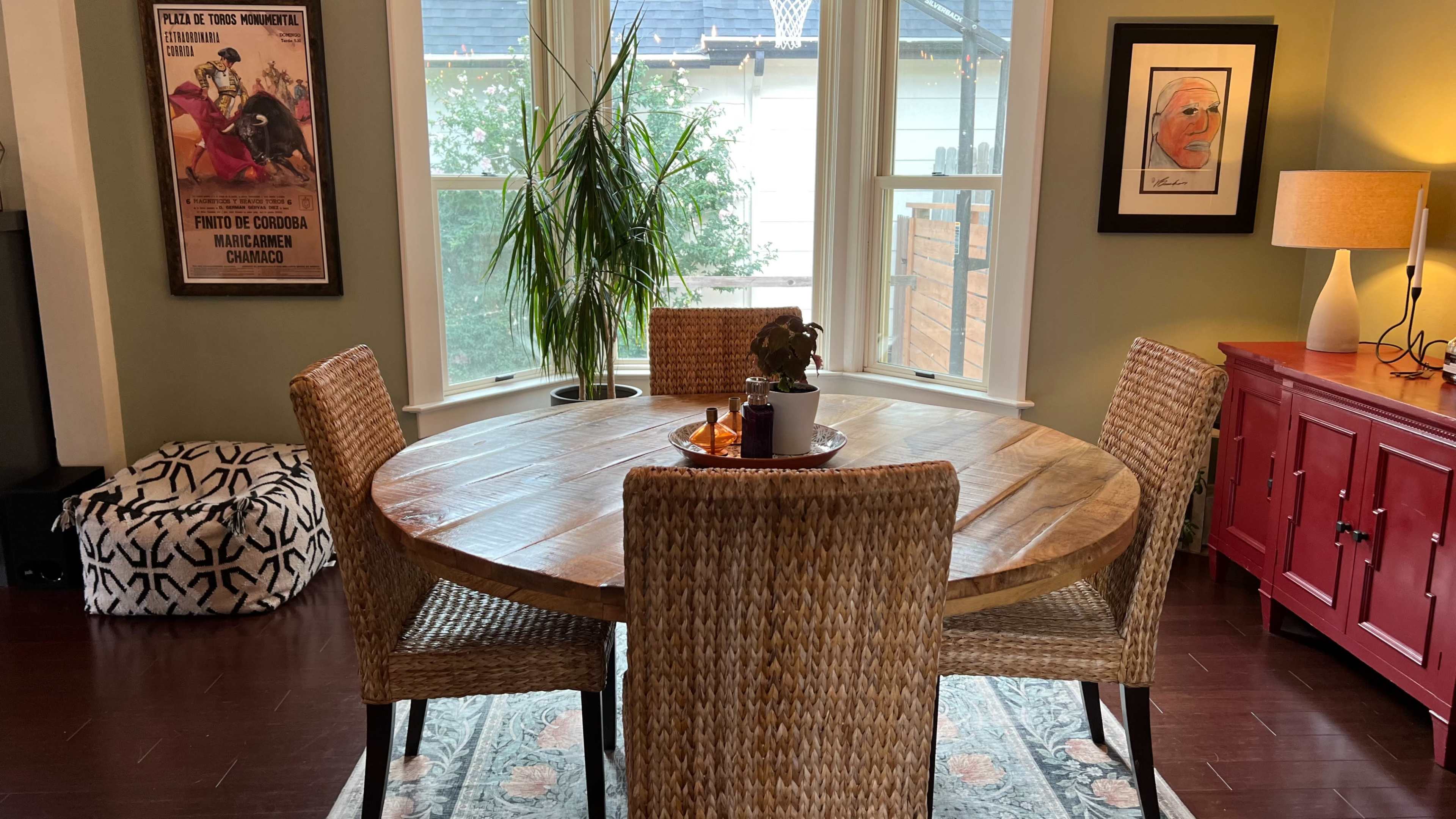 A round wooden dining table is set with a small plant and decorative items, surrounded by four woven chairs, near a window with natural light filtering in.