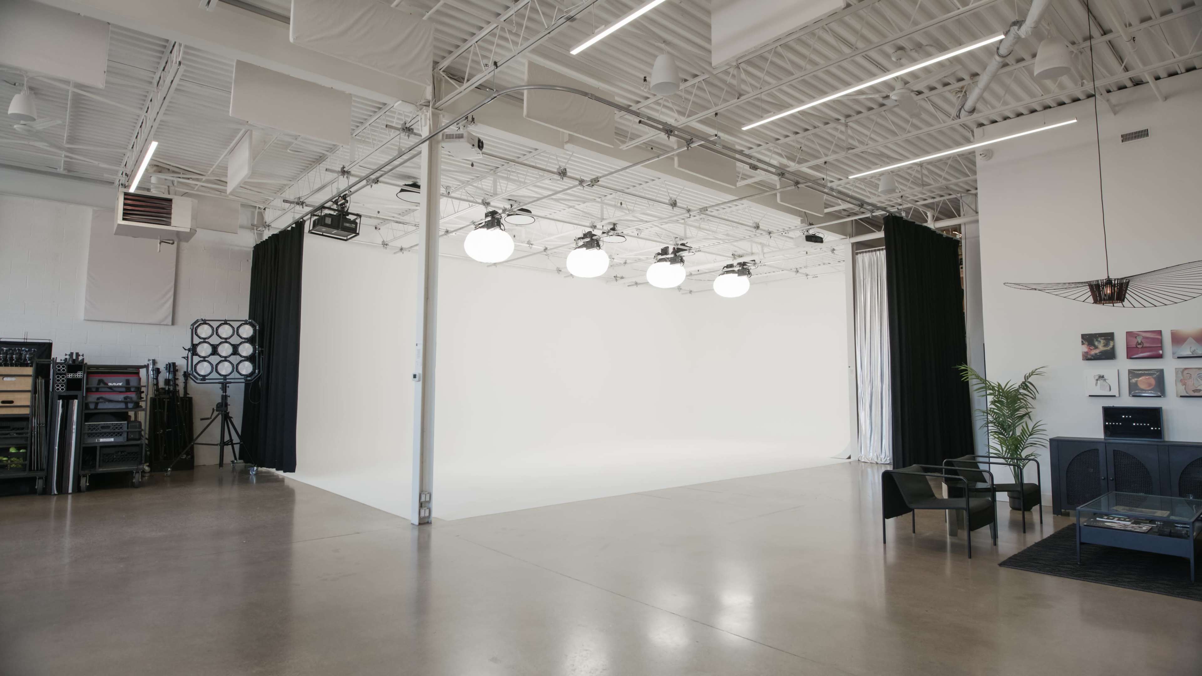 The image shows a spacious, well-lit studio with a blank white backdrop, ceiling lights, and minimalist furniture on one side.