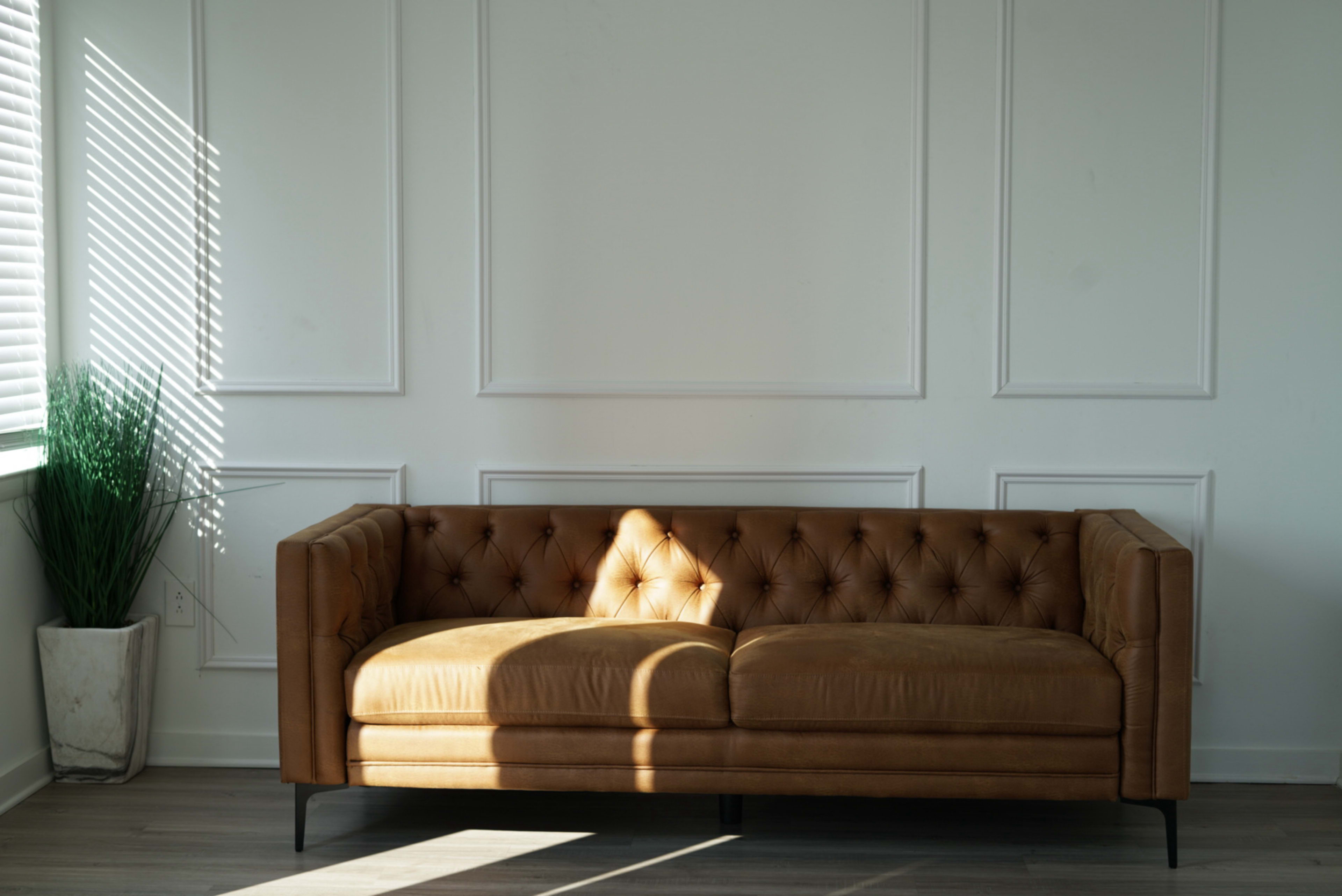 A brown tufted leather couch is positioned against a white wall with light streaming in from a nearby window, casting shadows on the floor.