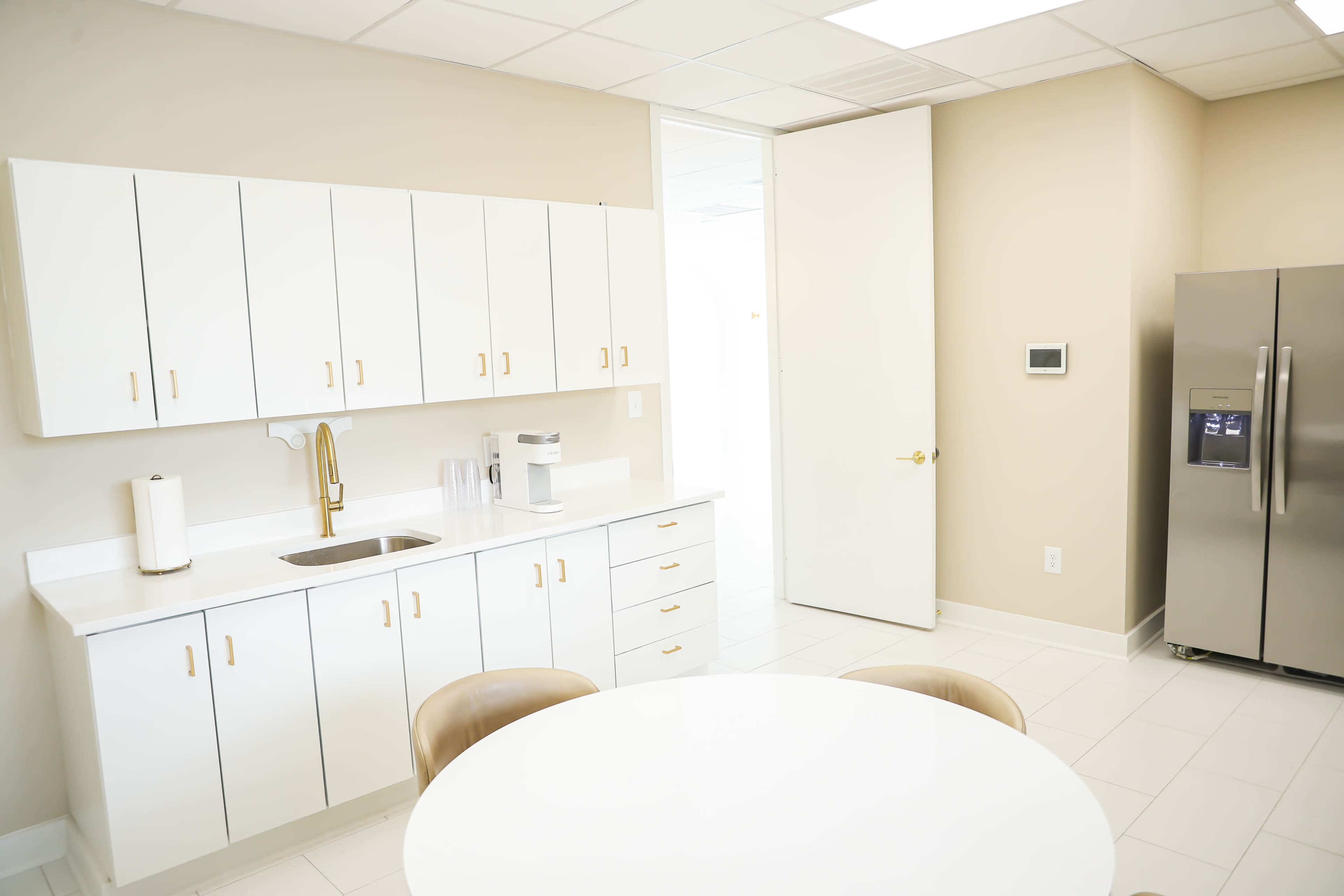 A bright kitchen features white cabinets, a sink, a coffee maker, and a round table with two chairs adjacent to a stainless steel refrigerator and a closed door.