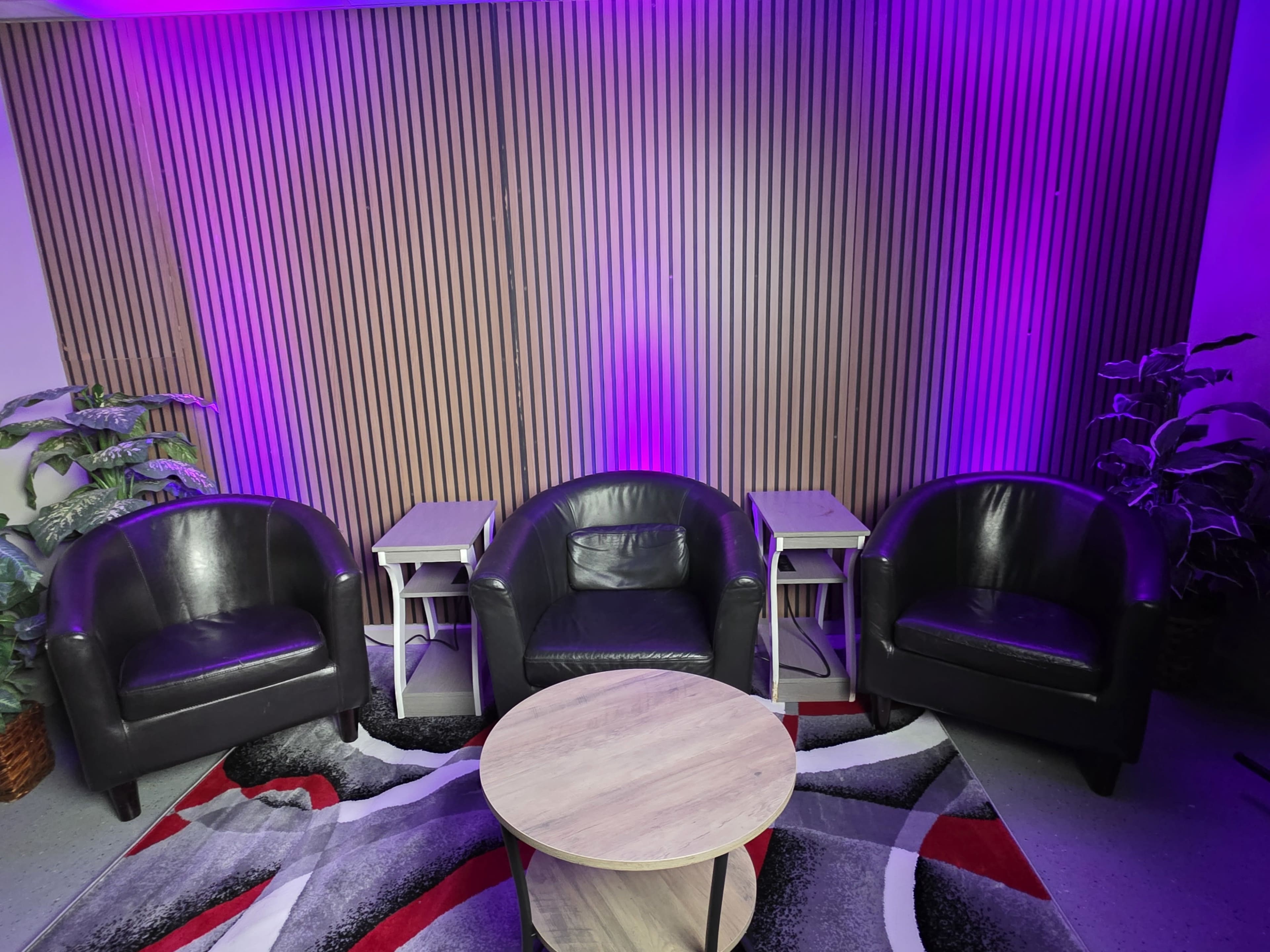 The image shows a seating area with three black leather chairs arranged around a circular wooden table, with purple lighting illuminating the wooden wall behind them.
