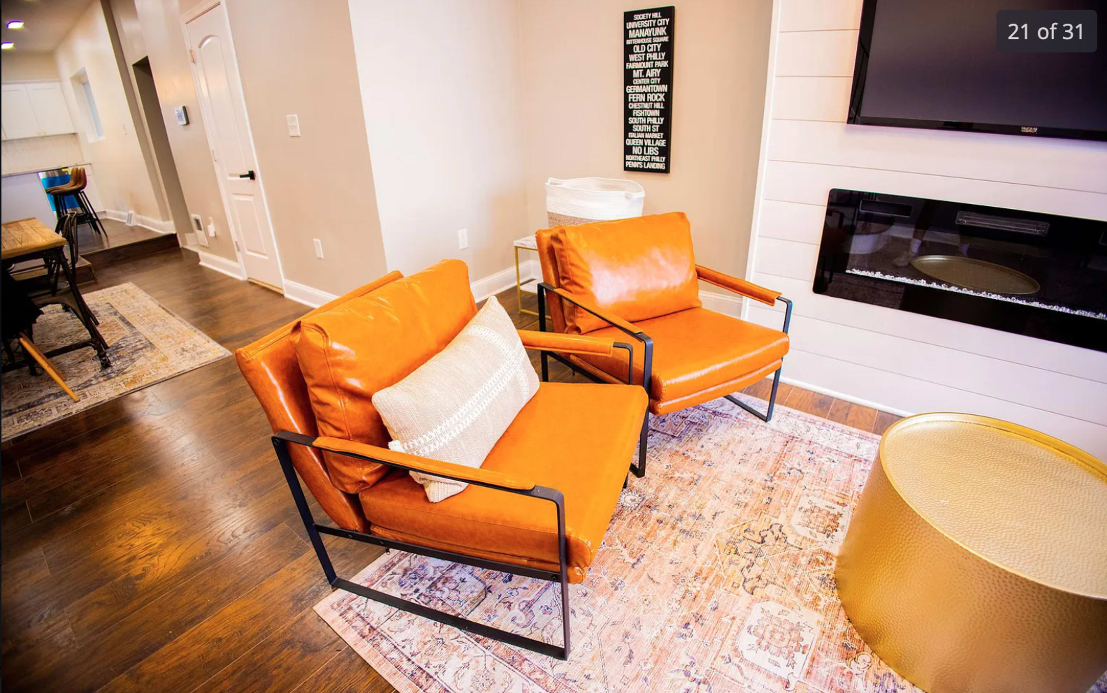 Two orange chairs are positioned on a rug in a modern living room, accompanied by a gold coffee table and a wall-mounted television.