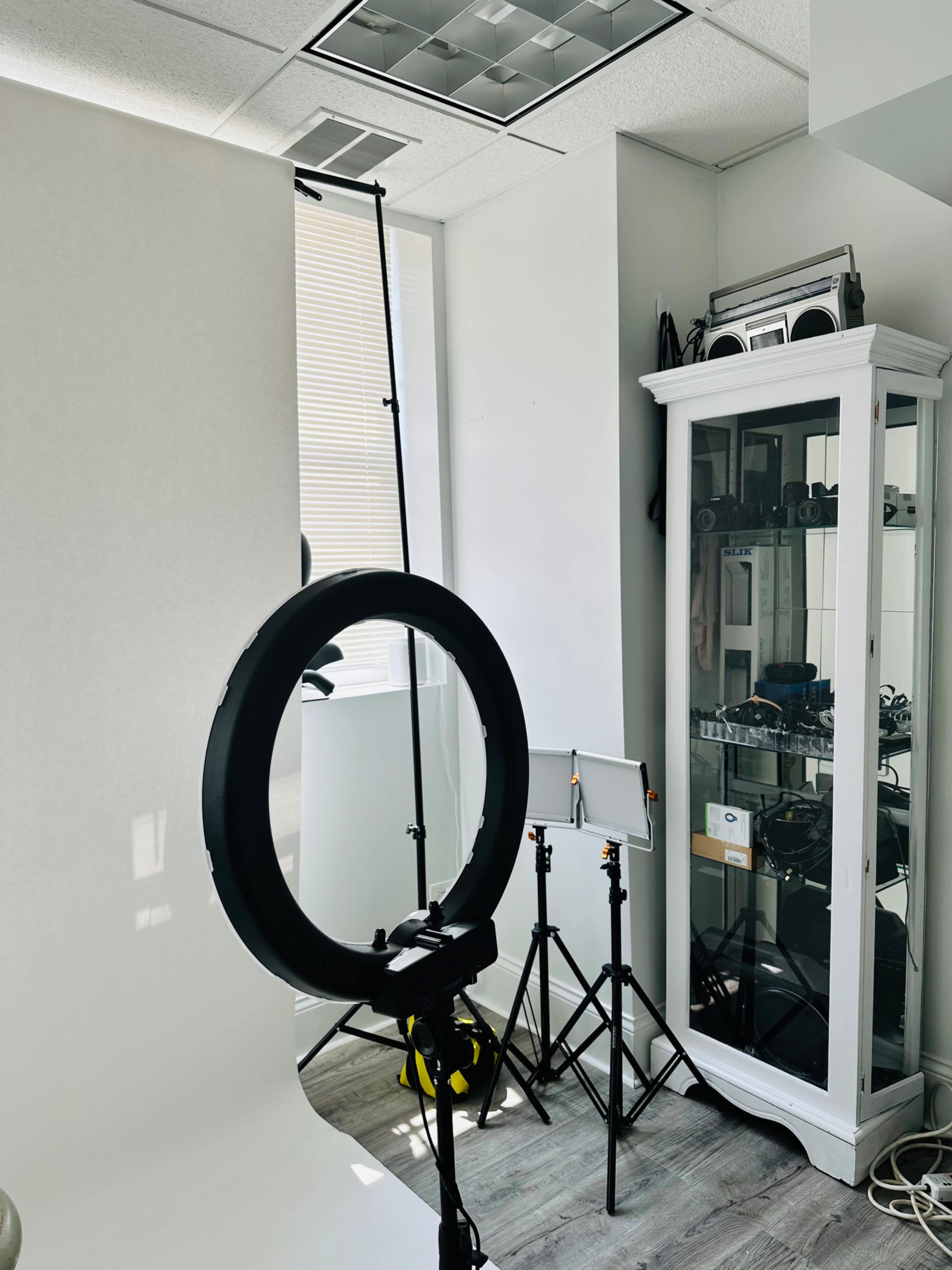 The image shows a well-lit photography studio featuring a ring light, a backdrop stand with a white backdrop, and a display case filled with photography equipment.