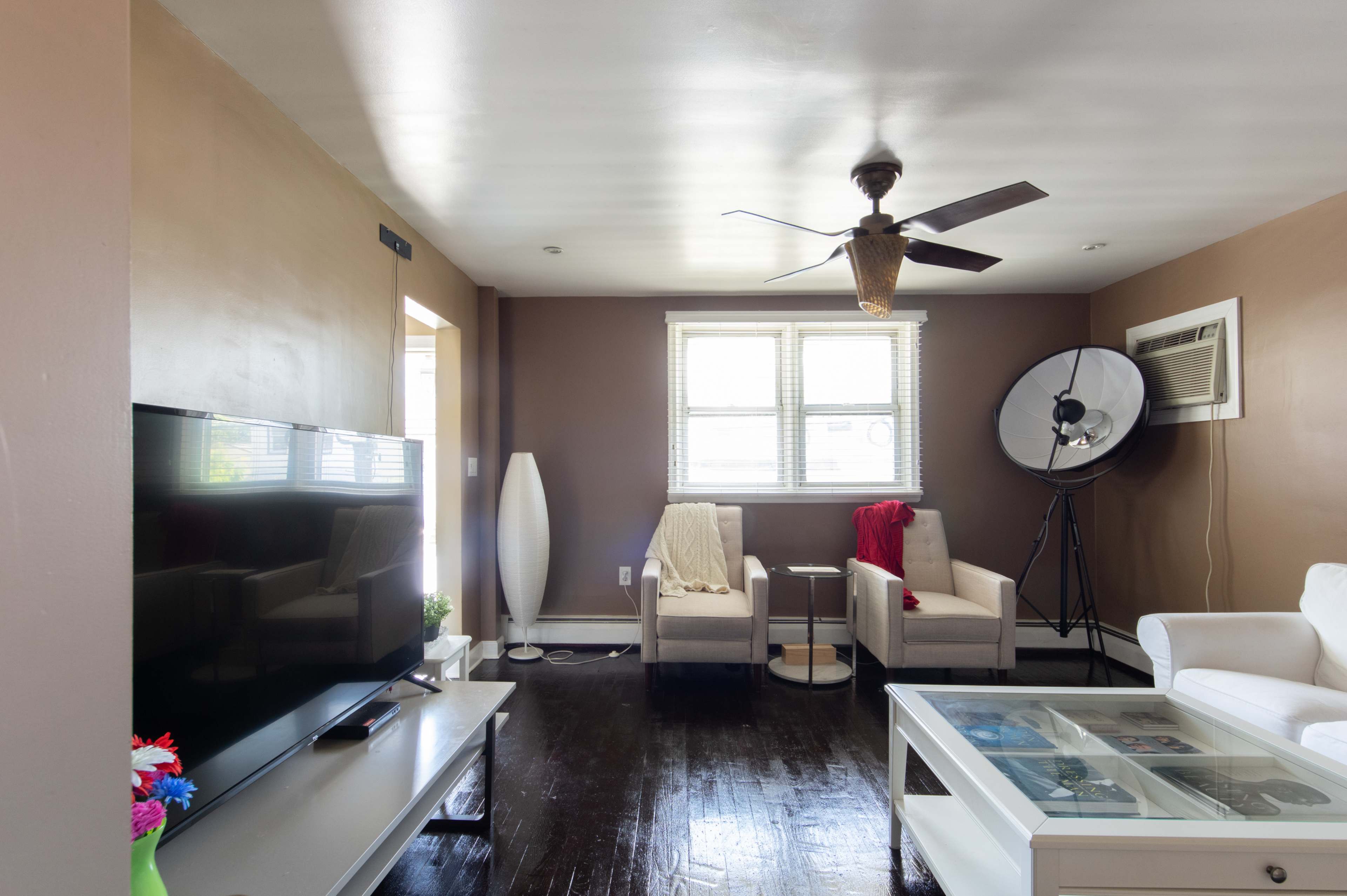 The living room features a large television, two armchairs, a fan, and a window with natural light, accompanied by a standing lamp and a photography light setup.