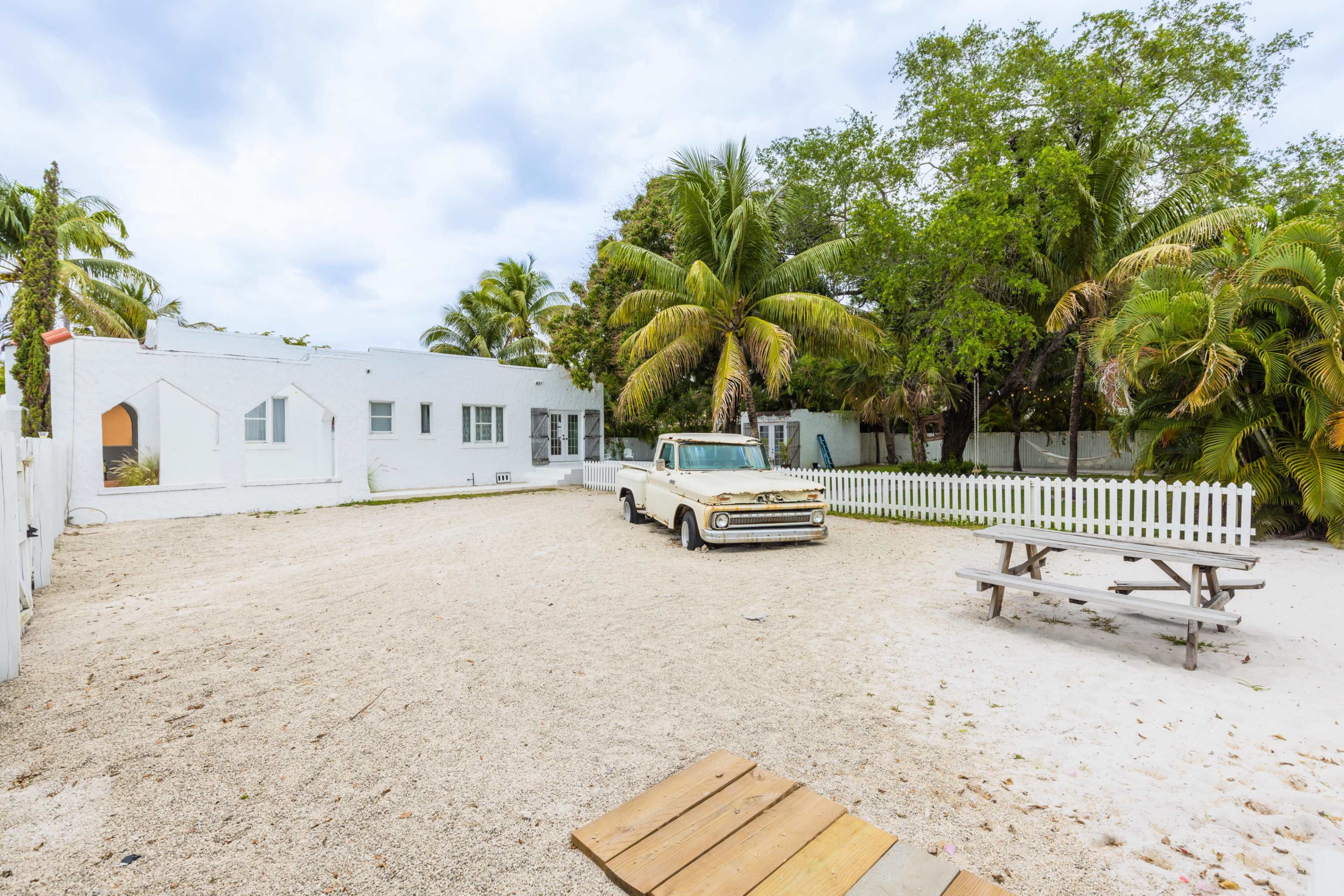 A light-colored truck is parked in a sandy yard surrounded by palm trees and a white picket fence, with a picnic table nearby.