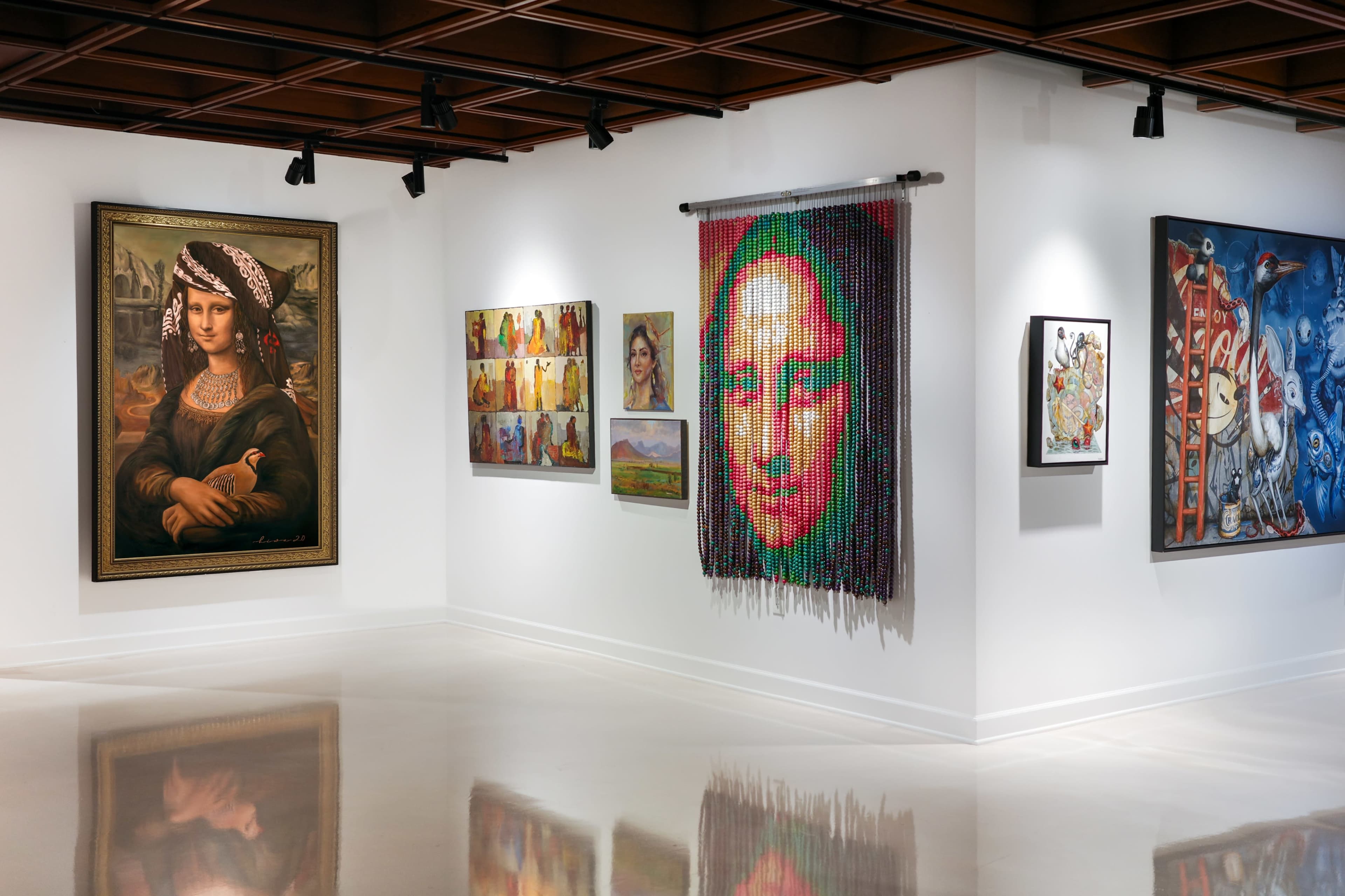The image shows an art gallery with various paintings and artworks displayed on white walls, including a large portrait and a colorful woven piece.