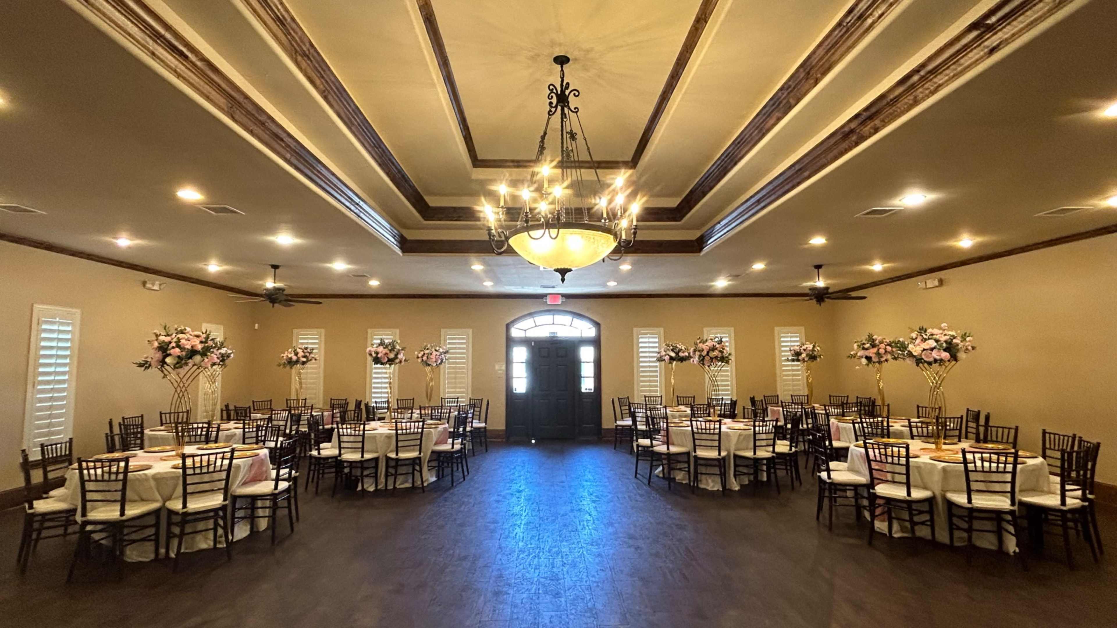 A spacious event venue set with round tables and floral arrangements, illuminated by chandeliers and natural light from the entrance.