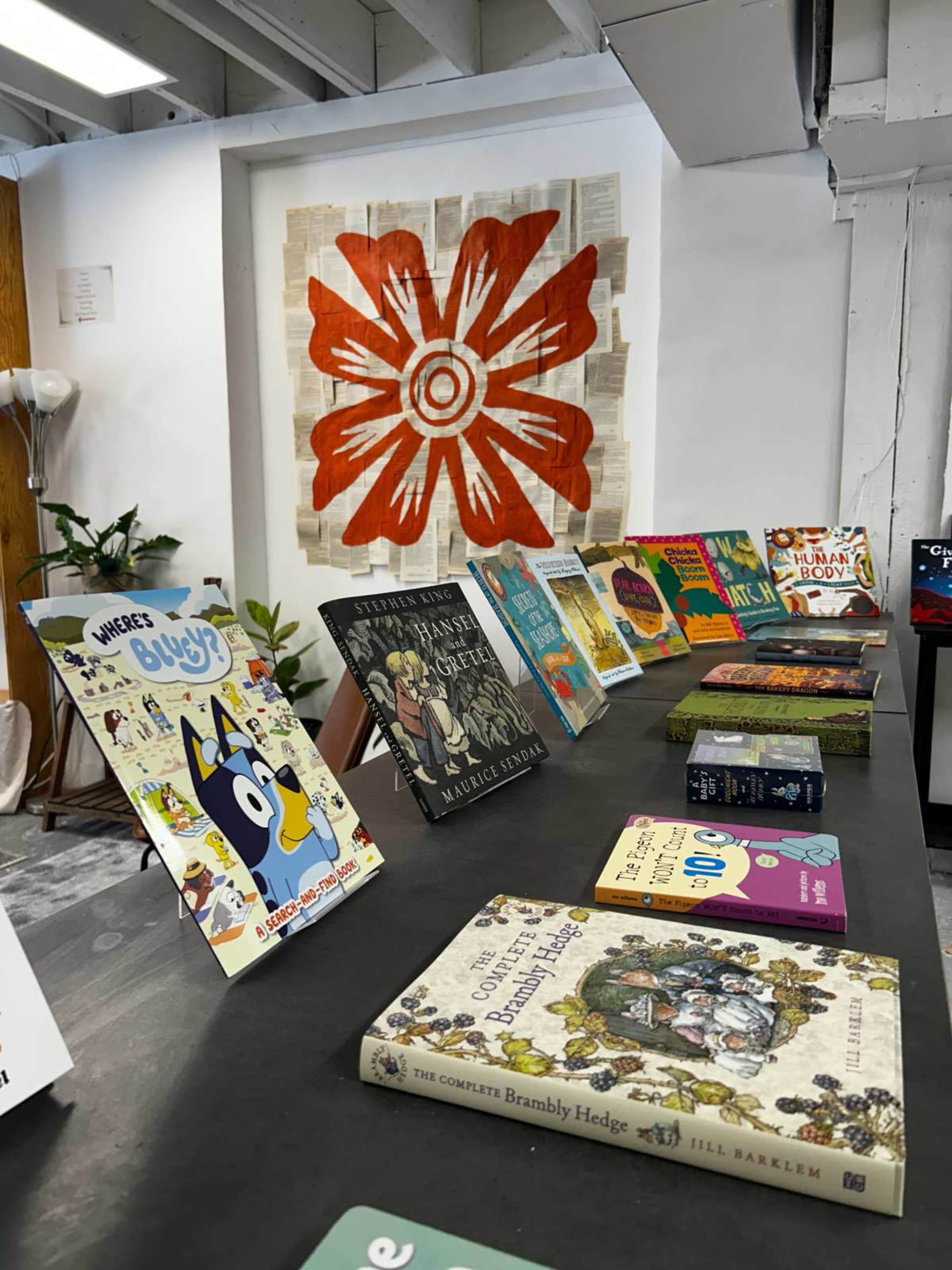 A row of colorful children's books is displayed on a table in front of a large red floral artwork on the wall.