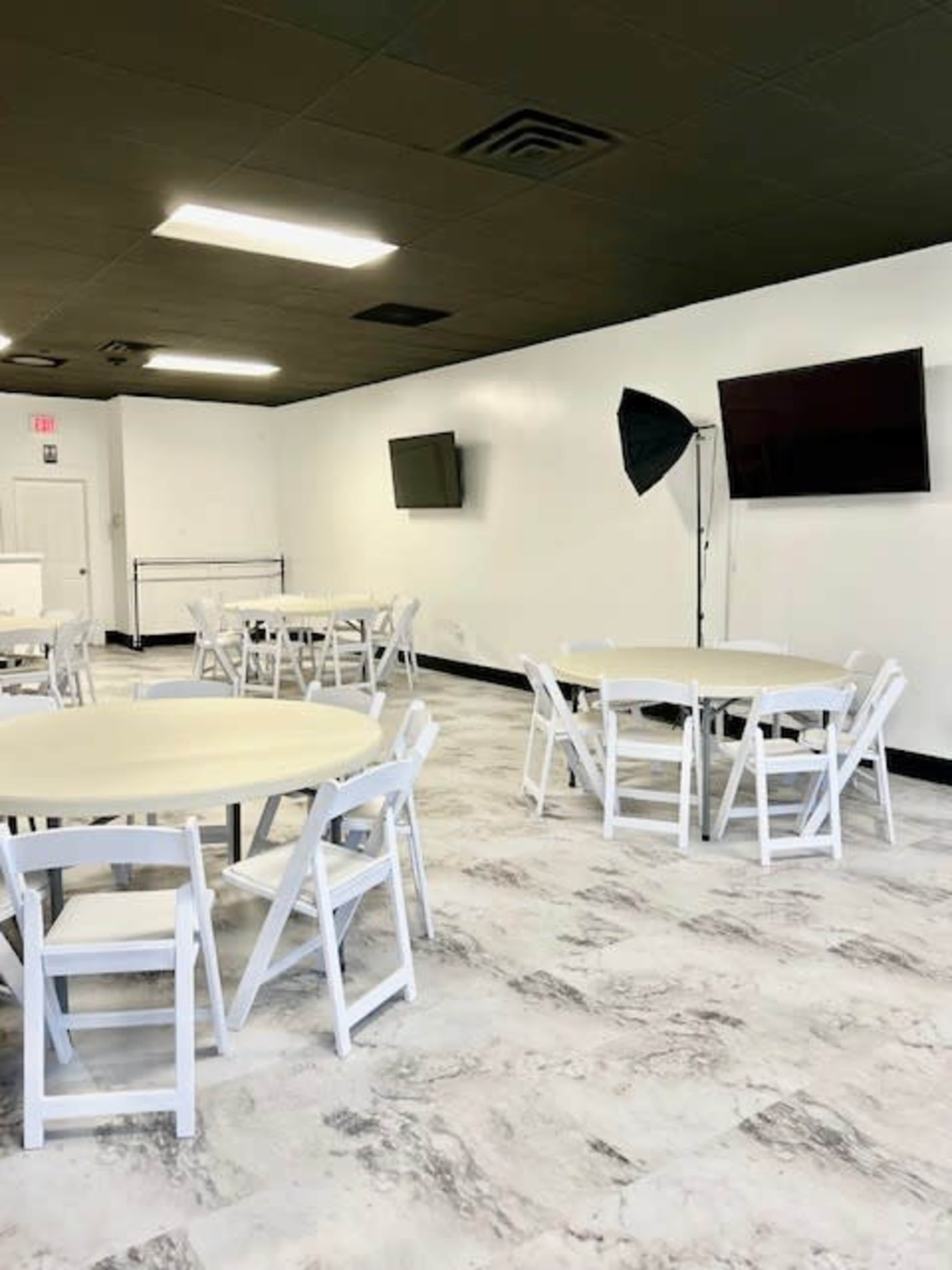 A spacious room with several round tables surrounded by white folding chairs, along with two wall-mounted televisions and studio lighting equipment.