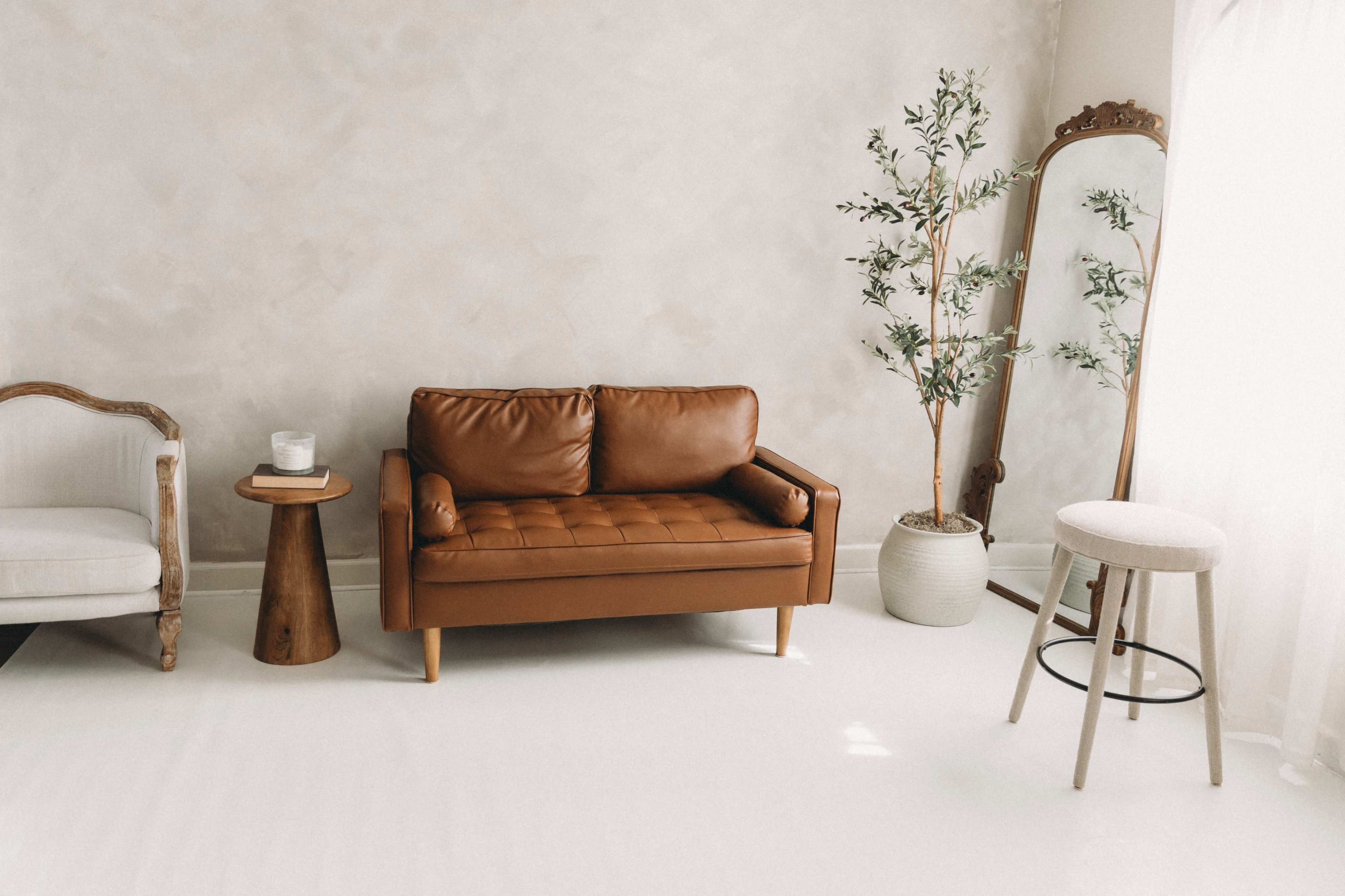 The image shows a minimalist living room with a brown leather sofa, a wooden side table, a tall plant, a full-length mirror, and a light-colored accent chair.