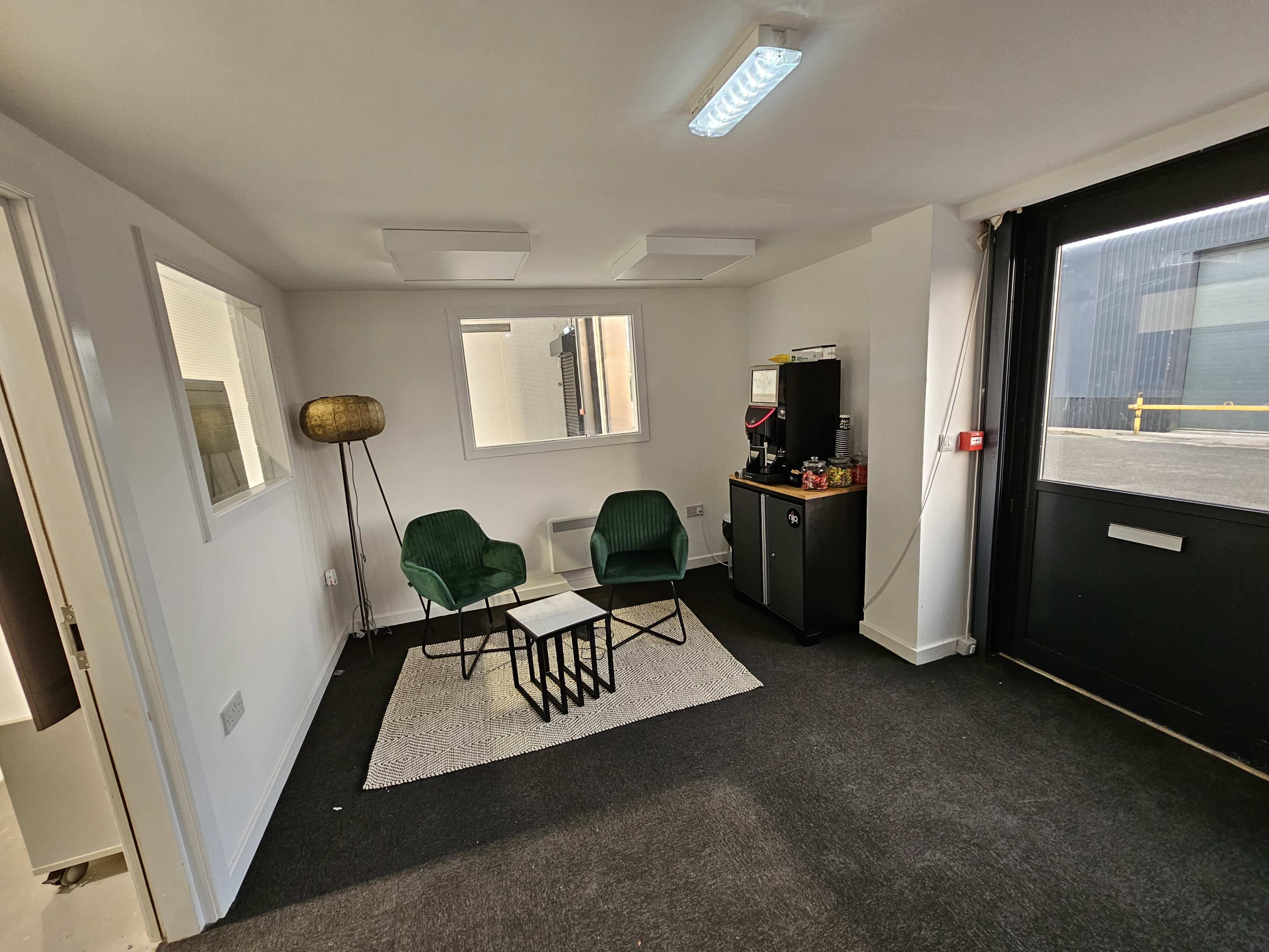 A small office space with two green chairs facing a coffee table on a rug, a refrigerator, and a large glass door leading to an exterior area.