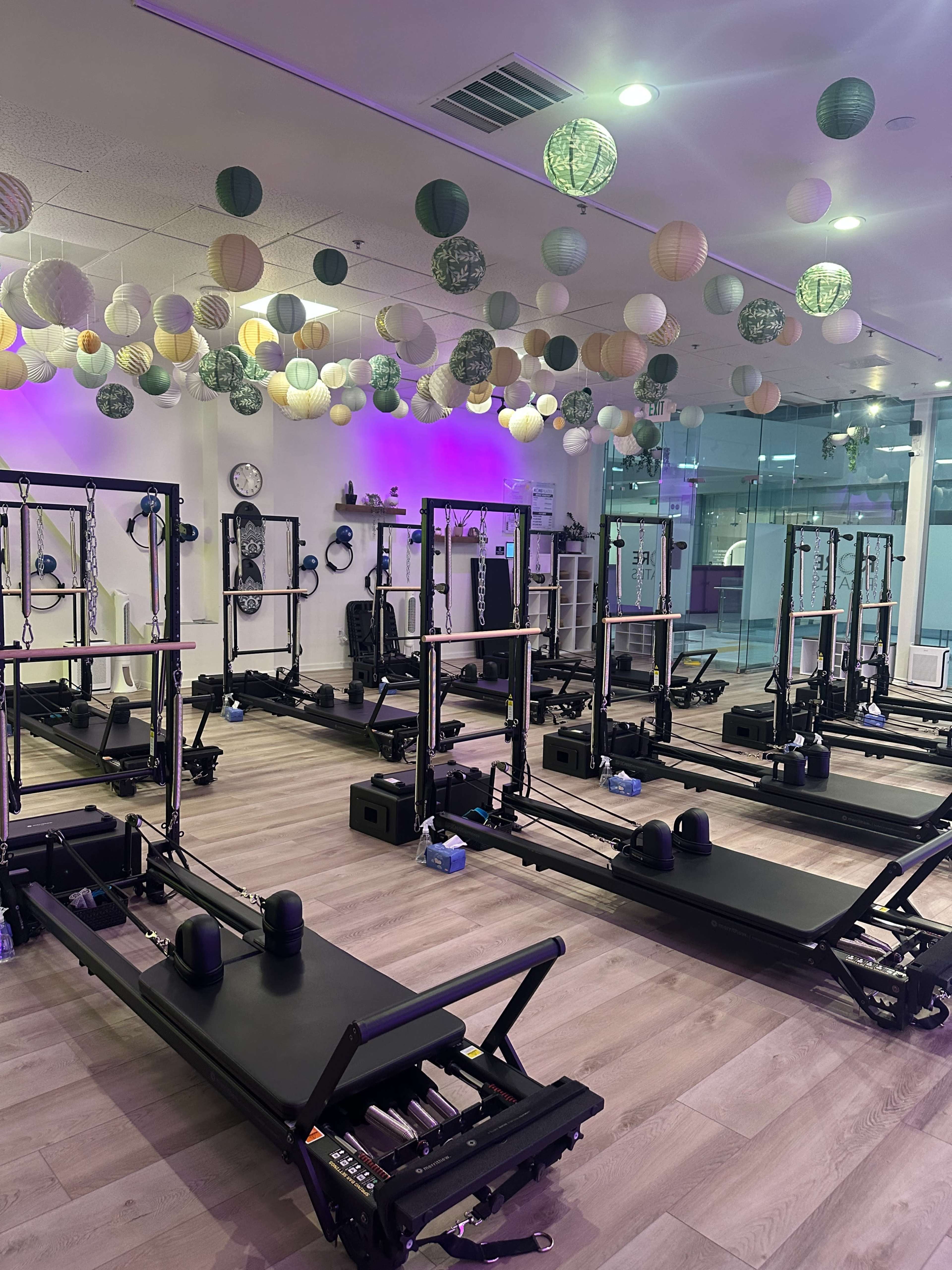The image shows a brightly lit fitness studio with several reformer Pilates machines set up in rows beneath decorative paper lanterns hanging from the ceiling.