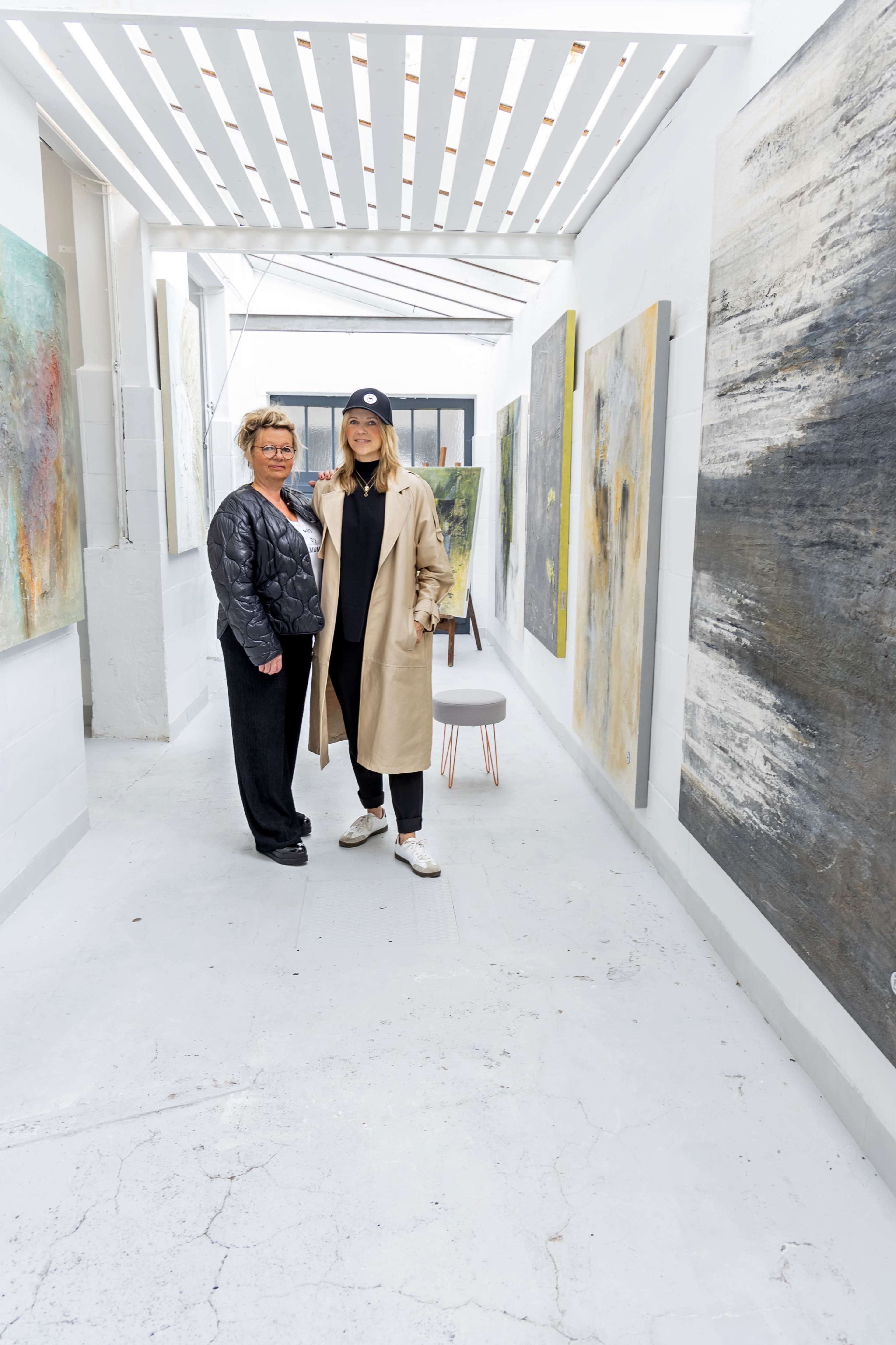 Two women stand in a brightly lit art gallery corridor featuring several abstract paintings on the walls.