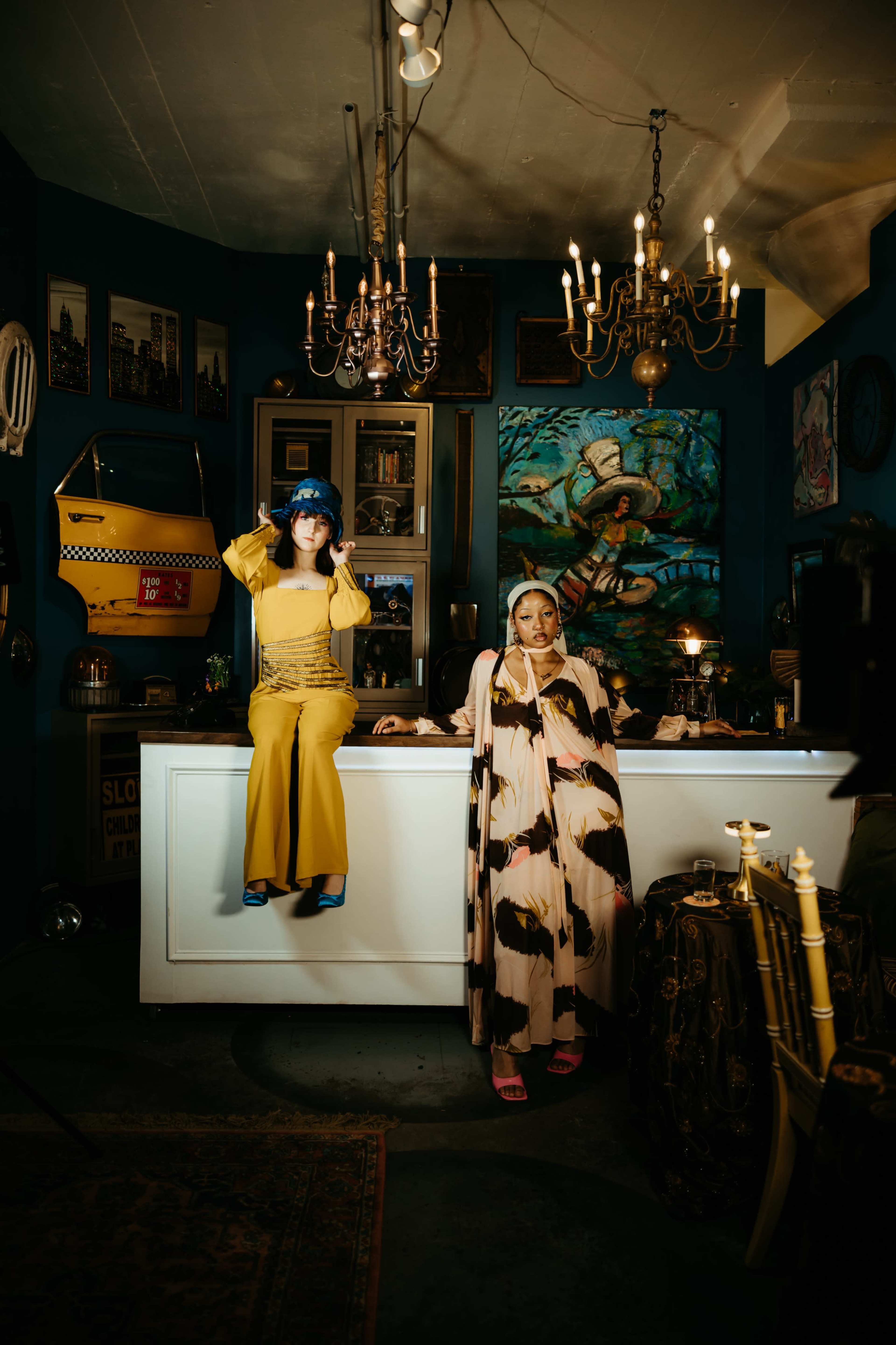 Two women dressed in vibrant outfits pose inside a stylish interior featuring art, chandeliers, and eclectic decor.