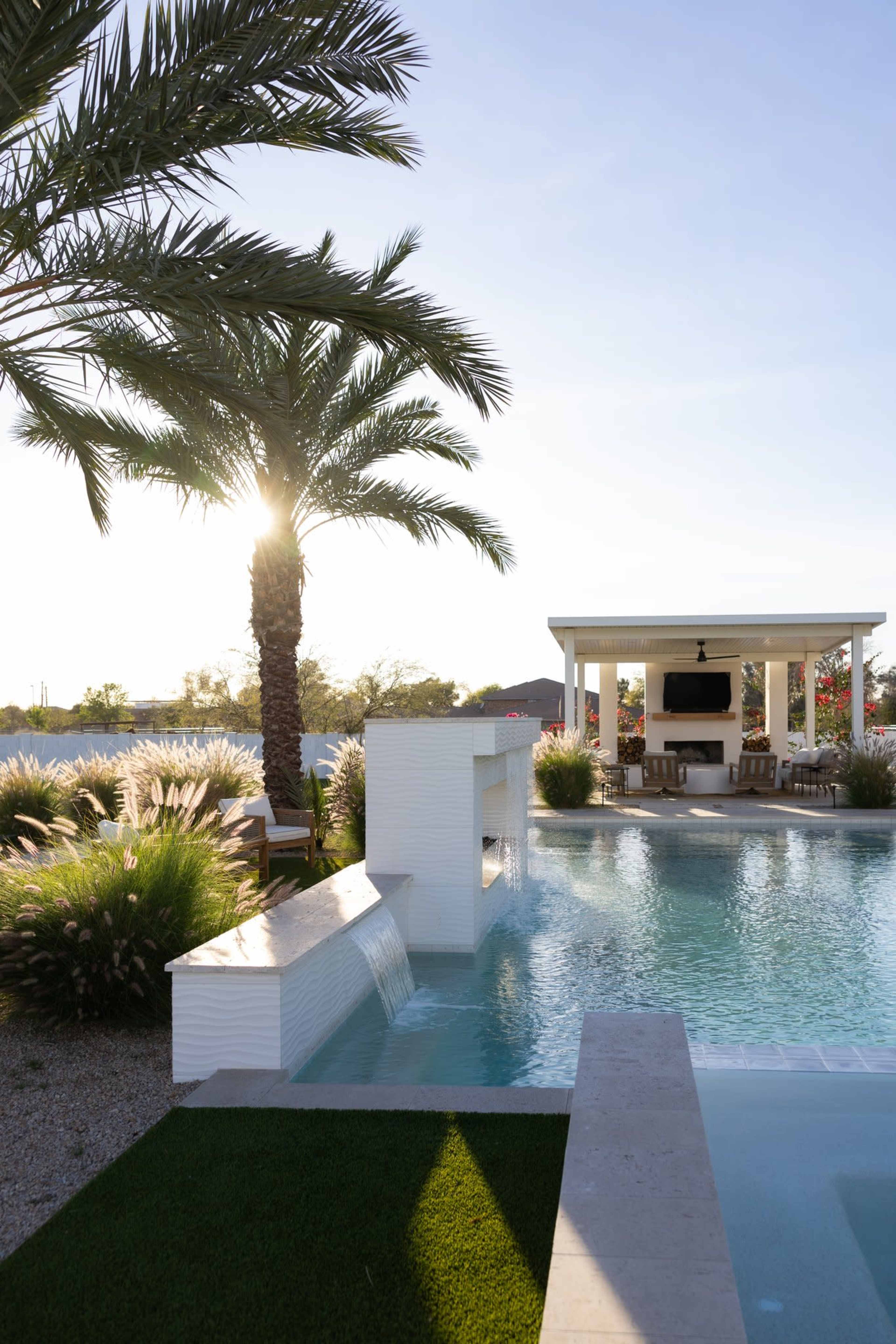 The image depicts a tranquil outdoor pool area featuring a cascading water feature, palm trees, and a covered seating area with a television.