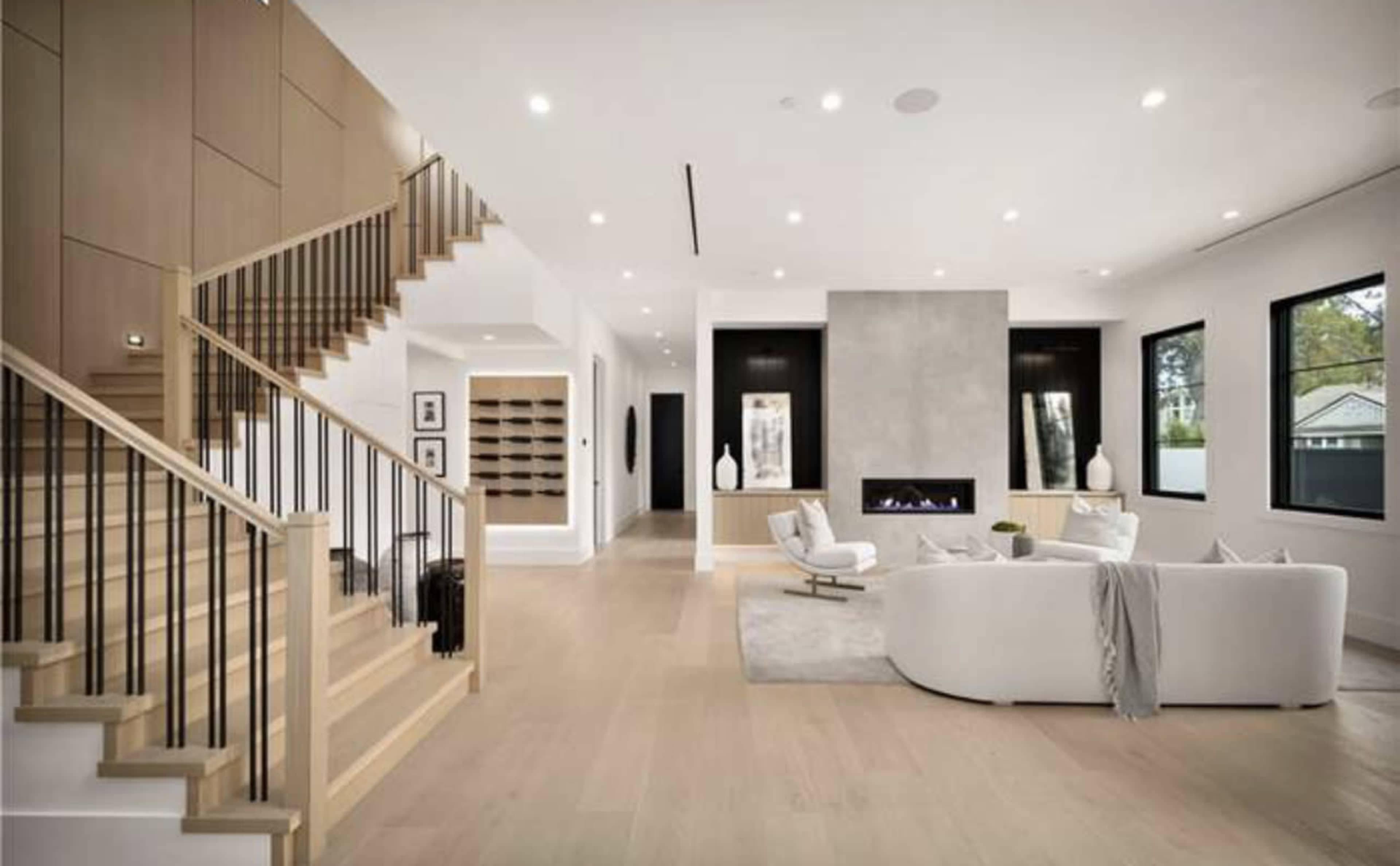 A modern living room with a white sofa, a fireplace, and a staircase in a spacious area featuring large windows and light wood flooring.