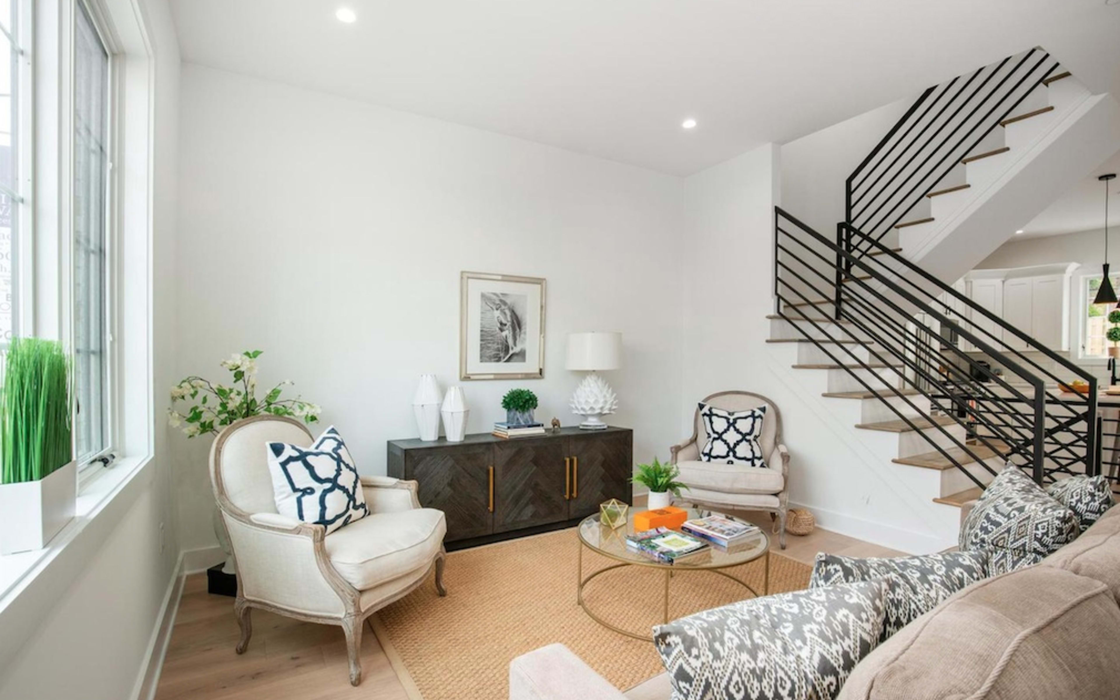 A bright living room features two upholstered chairs, a round coffee table, and a staircase leading to an upper level.