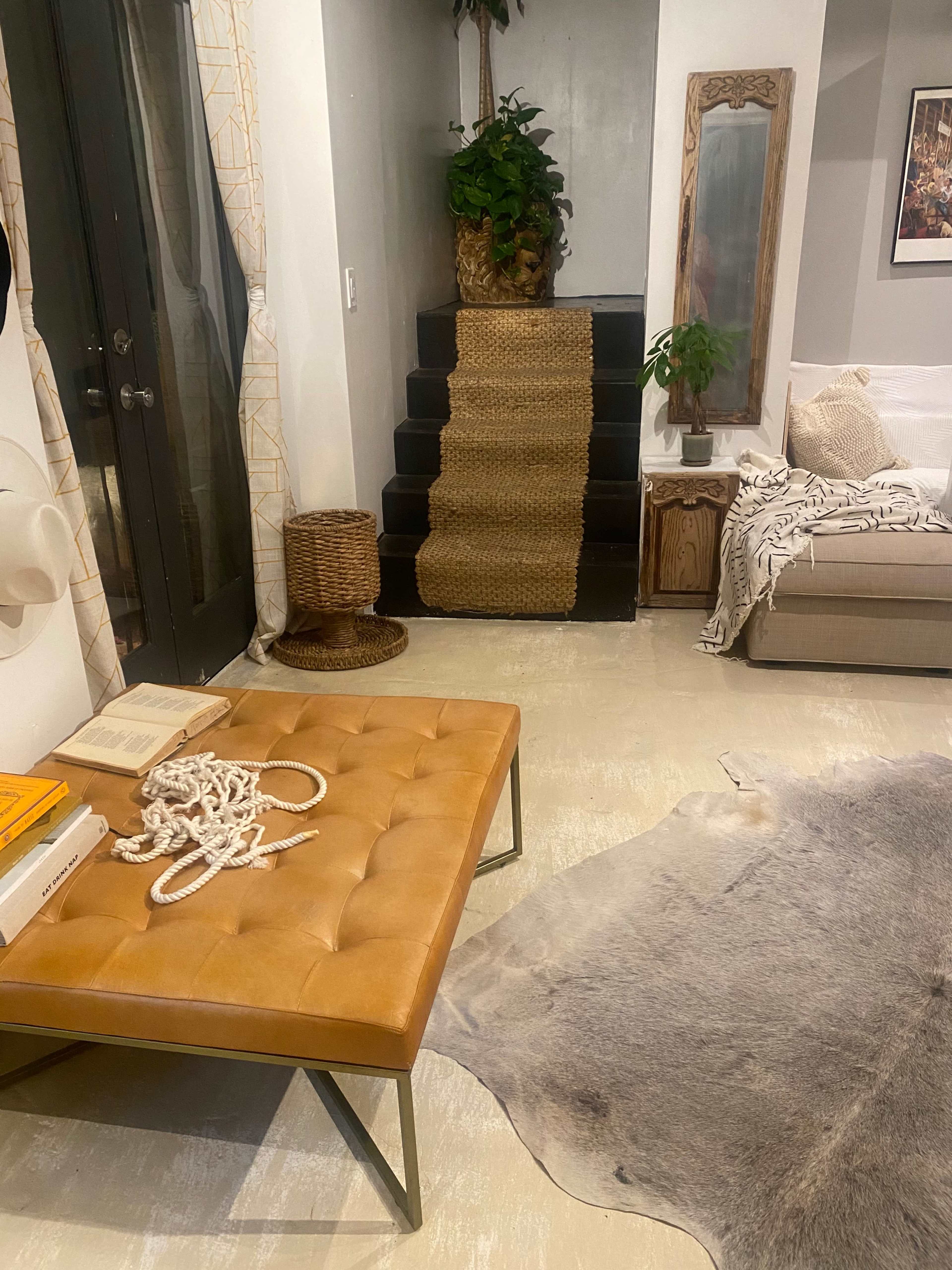 The image shows a living room with a tan leather ottoman, a staircase covered in a woven rug, a potted plant, and a sofa along the wall.