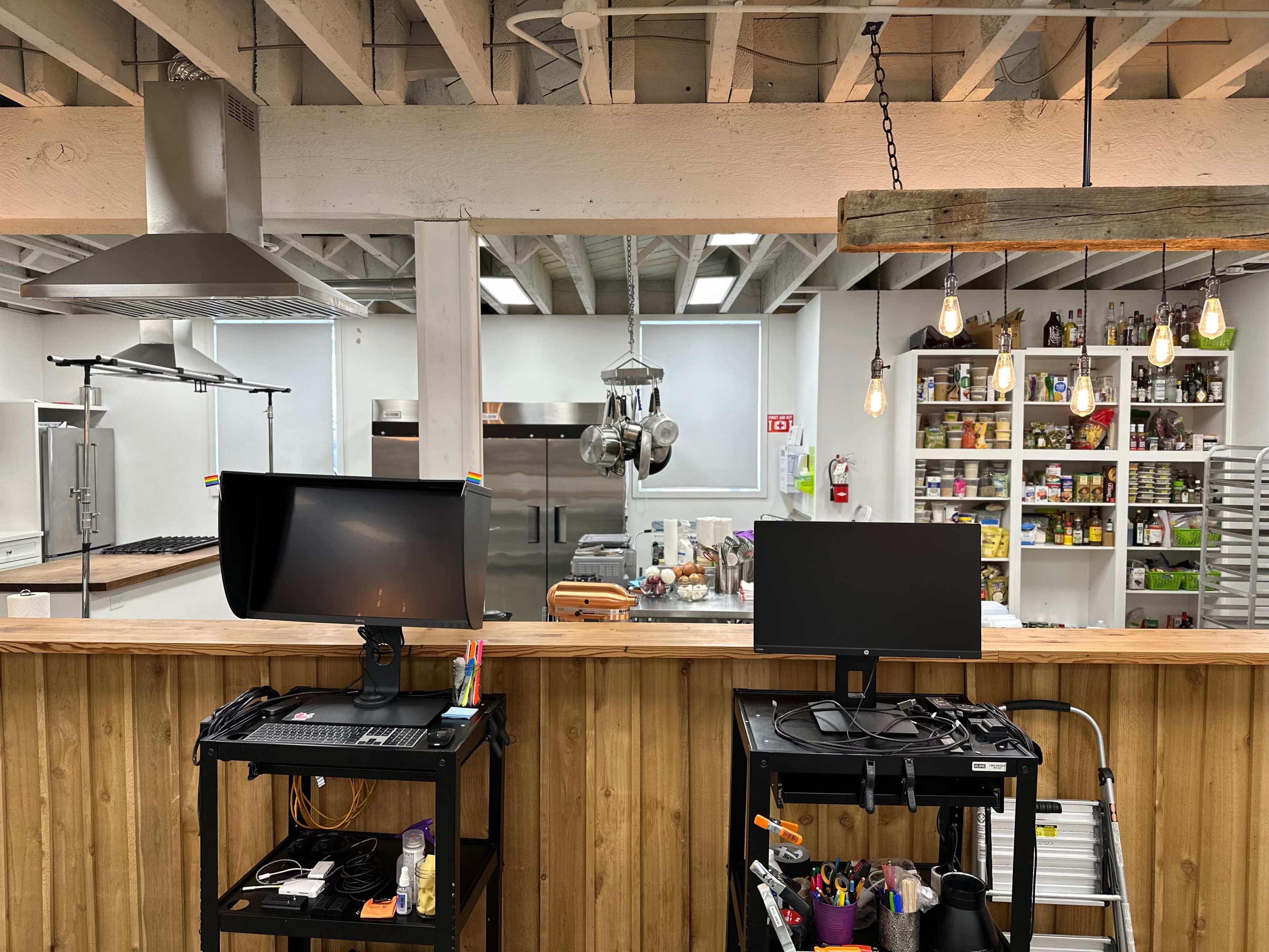 A kitchen space with two computer monitors on a wooden counter, while shelves in the background display various food items and utensils.