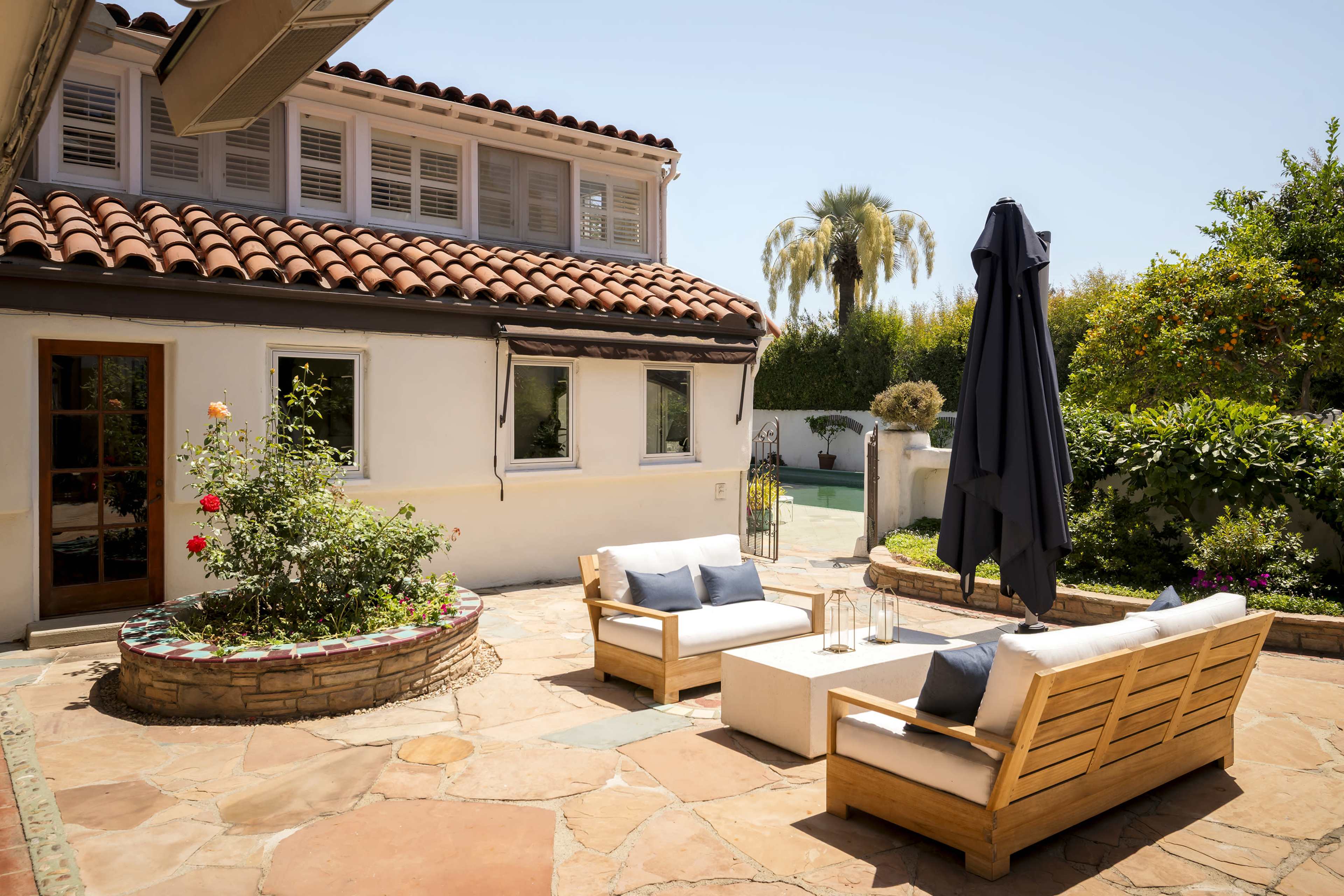 The image shows a sunlit outdoor patio with two wooden sofas, a coffee table, and an umbrella, surrounded by greenery and a stone pathway.