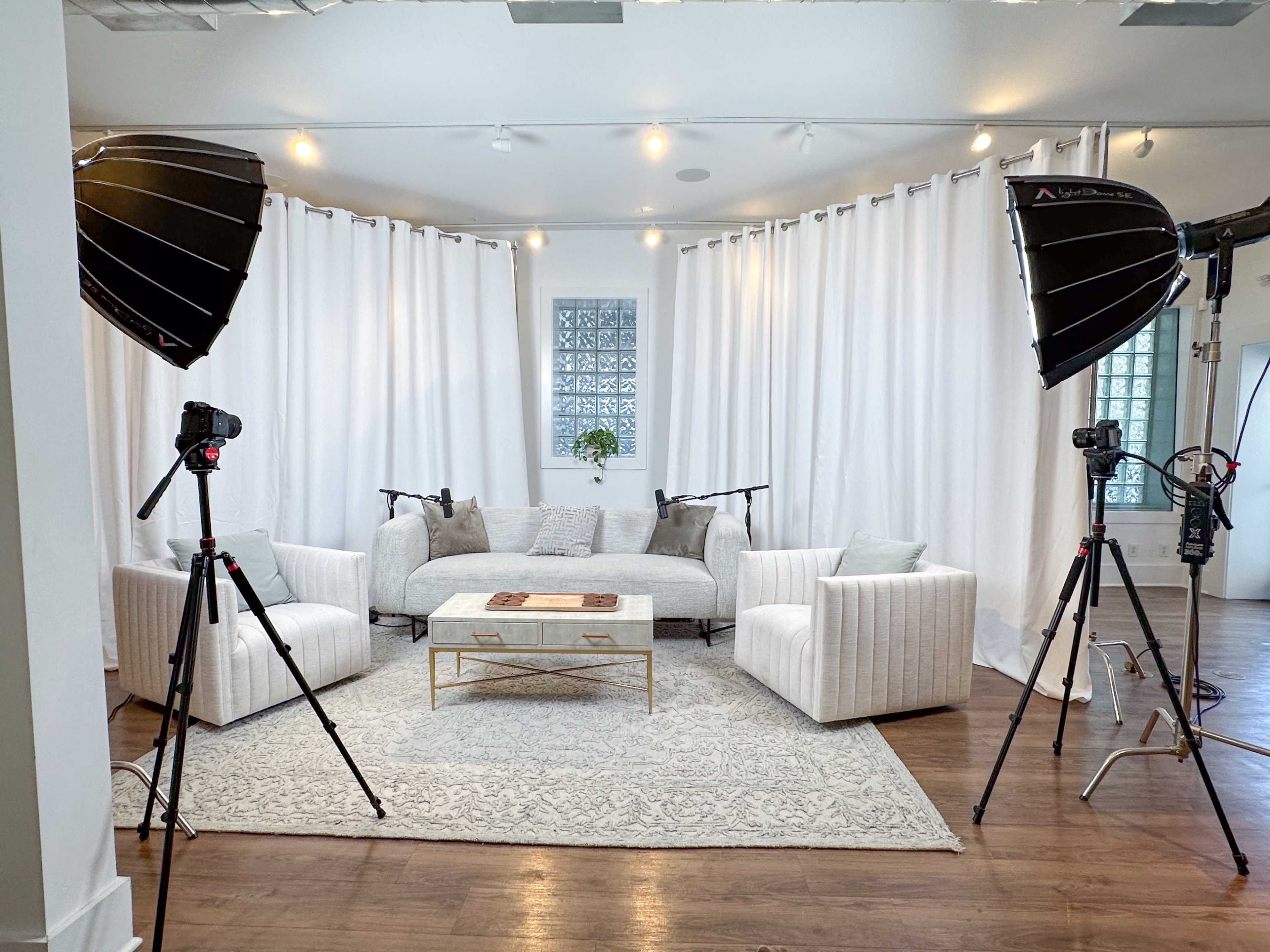 The scene features a bright studio space with a light-colored couch, two armchairs, a coffee table, and soft white curtains, all framed by photography lighting equipment on tripods.