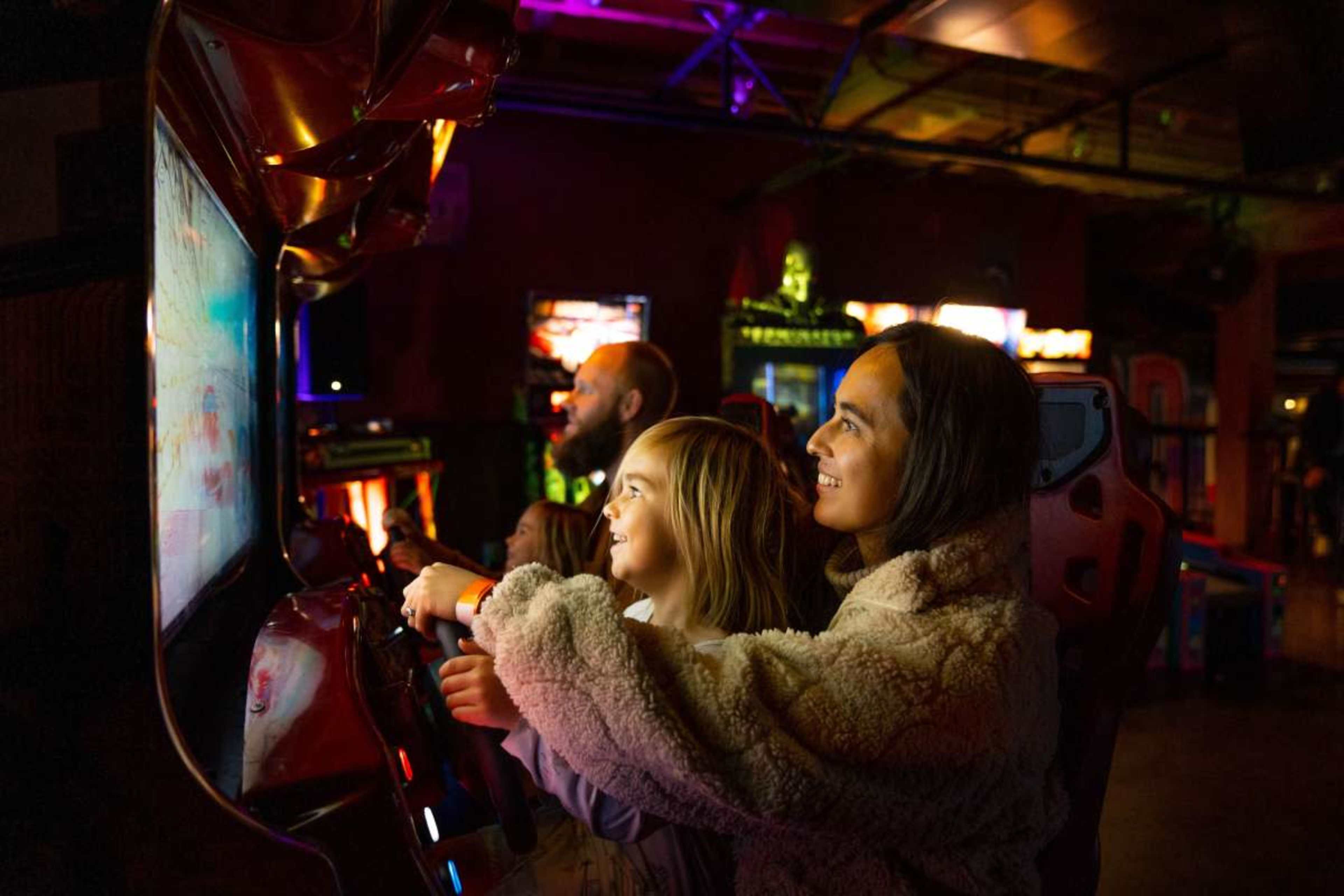 Two children play an arcade racing game while an adult observes in a brightly lit game room.