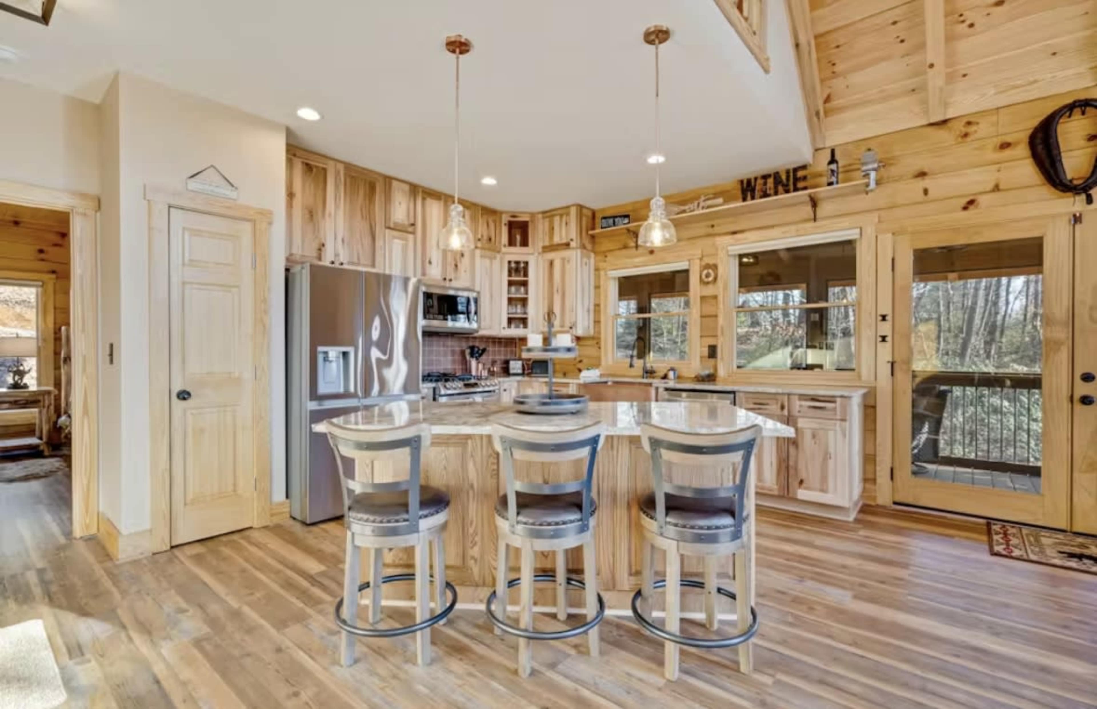The image shows a well-lit, wooden kitchen featuring a central island with three bar stools, modern appliances, and large windows overlooking a wooded area.